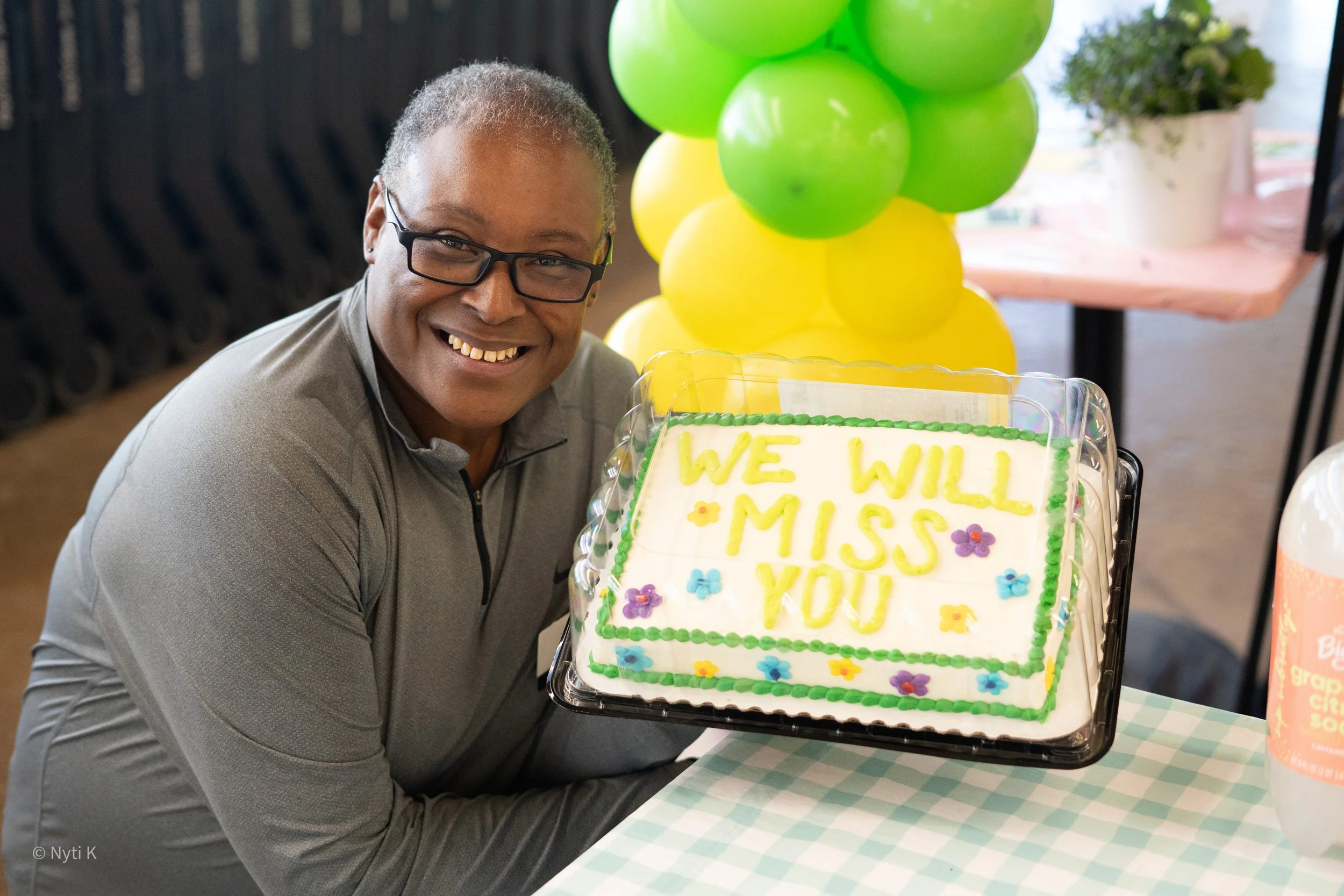 A smiling man with glasses is sitting at a table with a decorated cake that reads 'We will miss you.' There are green and yellow balloons in the background, and the table has a blue and white checkered tablecloth.