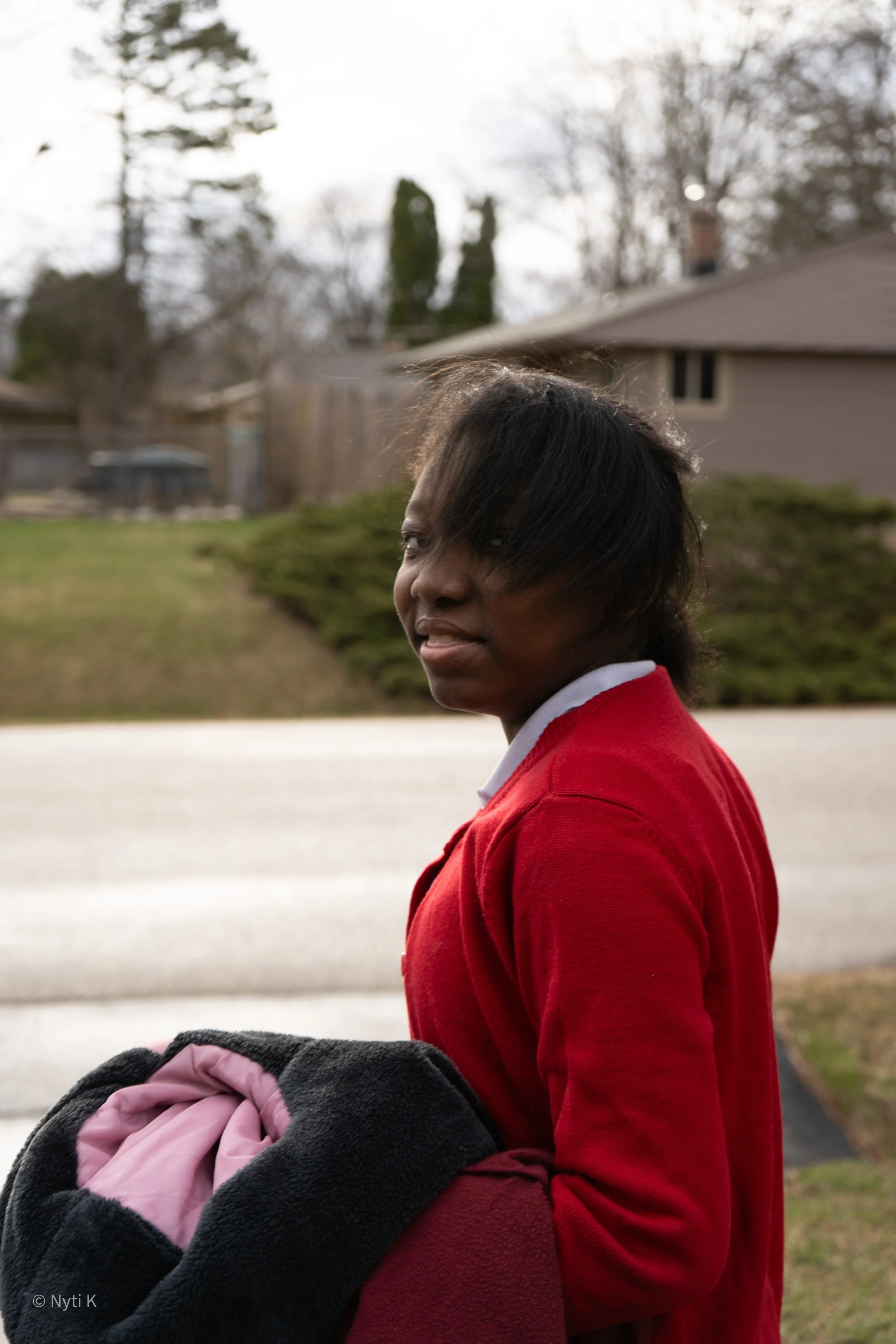 A woman with dark hair wearing a red sweater stands outdoors in a residential area during daytime, holding a jacket or blanket. The background includes a house with a chimney, trees, and shrubs.