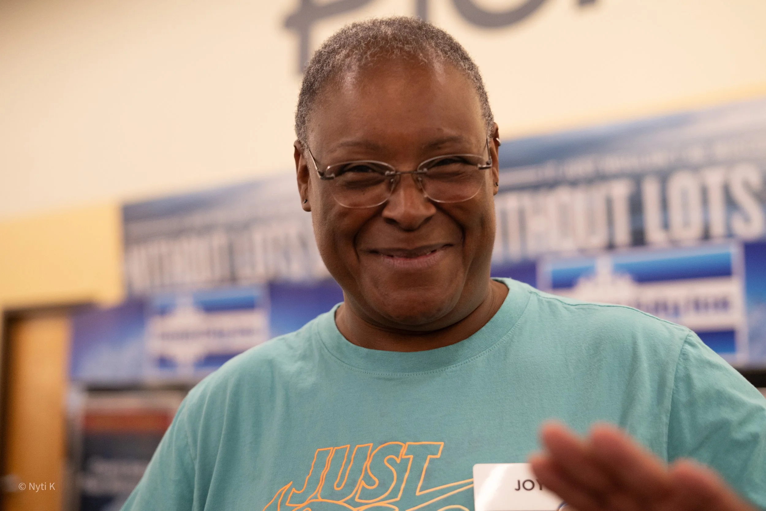 Smiling woman with short gray hair and glasses, wearing a teal shirt, in a store or office setting.