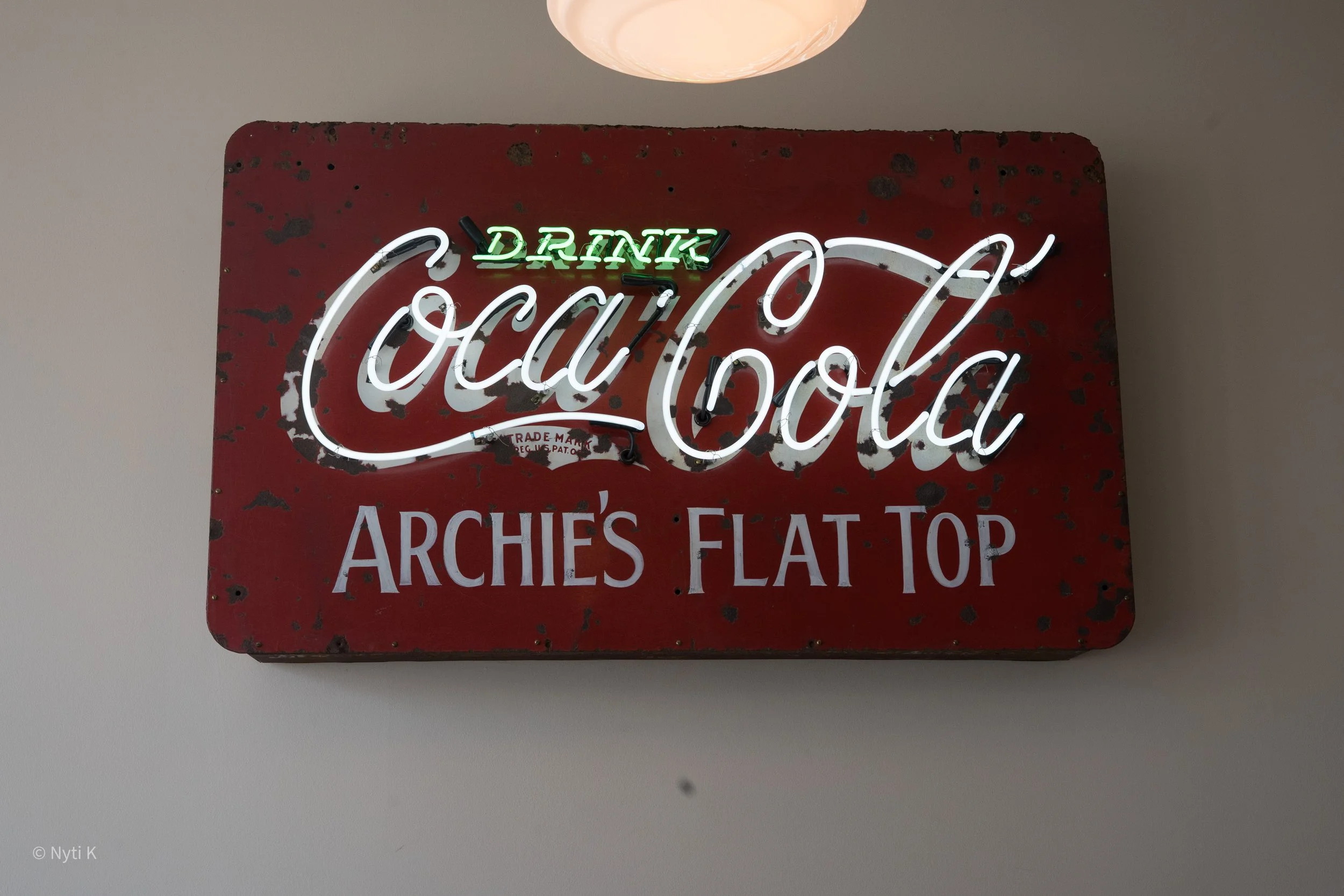 A vintage, rusted red metal sign with neon lights displaying the word "Coca-Cola" in cursive, along with the words "Drink" in green above it, and additional unreadable text at the bottom.