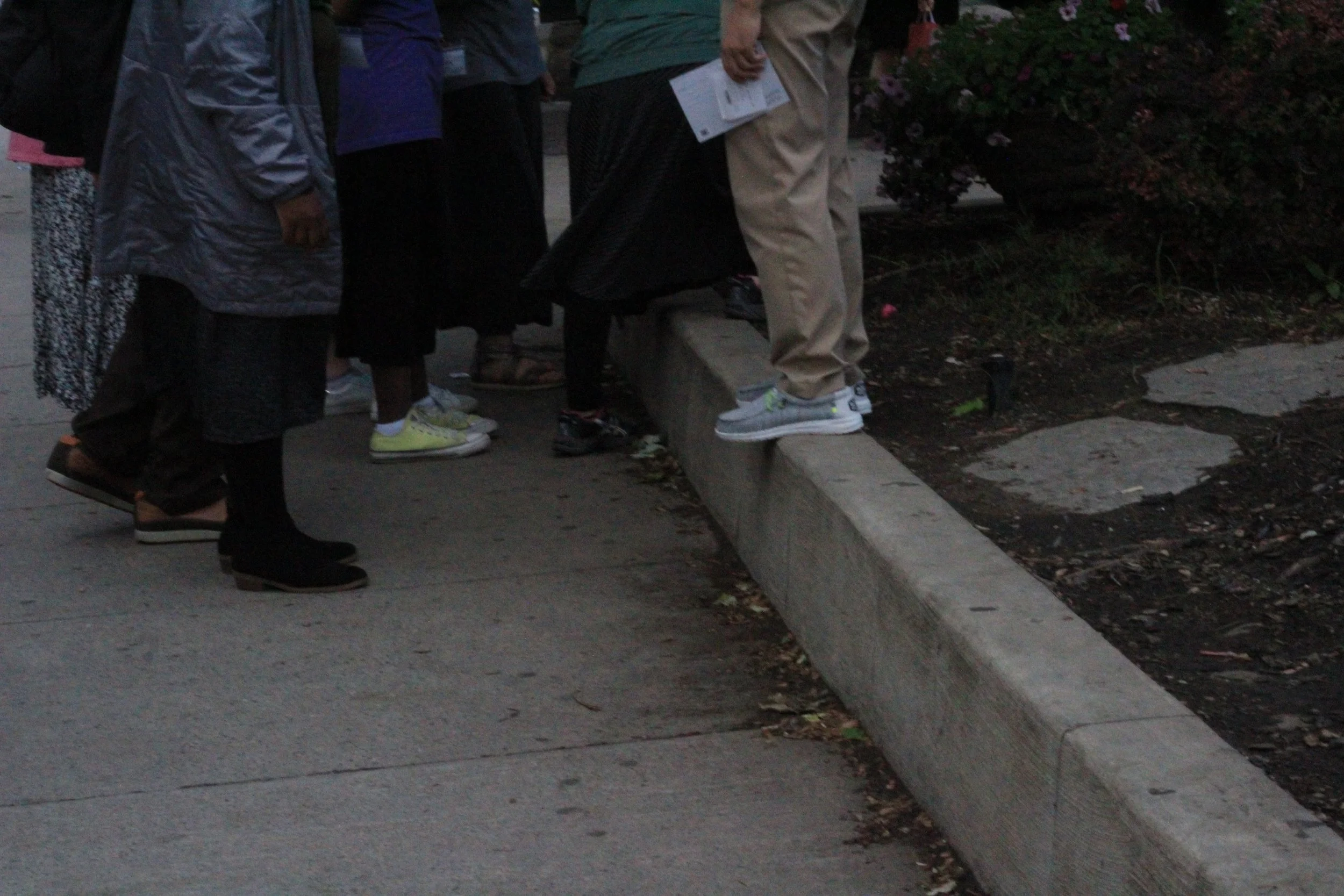 Close-up of a line of people waiting on a sidewalk, wearing casual clothing and sneakers, some holding papers. The sidewalk has a curb and grassy area with bushes.