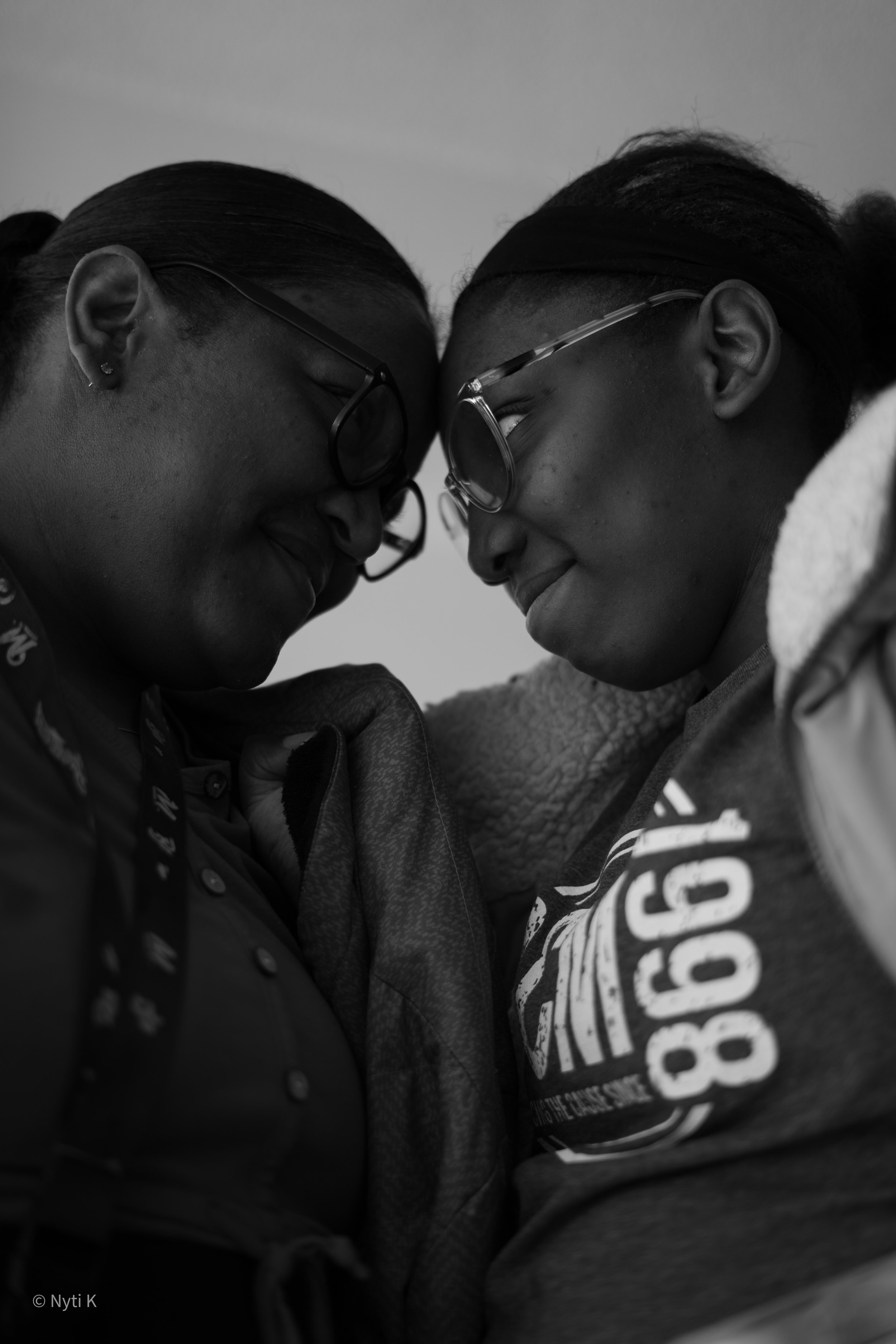 Two young women with glasses pressing foreheads together, smiling, in an intimate moment. Black and white photo, close-up of faces.