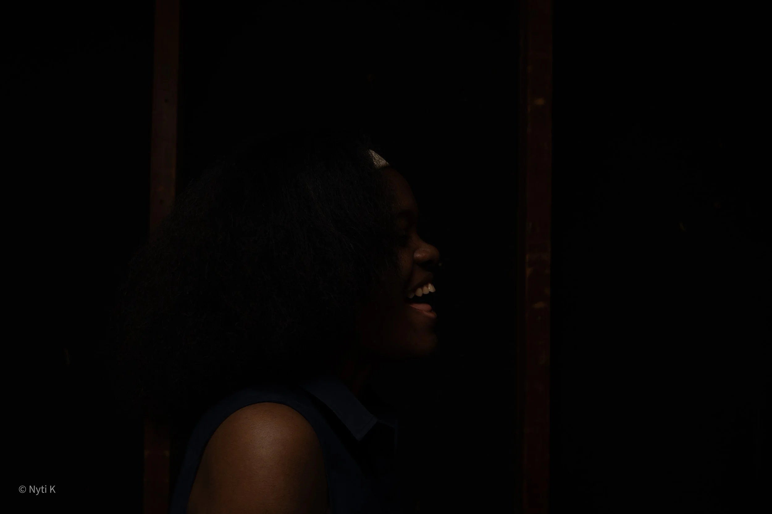 A woman with short hair smiling and showing her teeth, wearing a sleeveless top, captured in profile against a dark background.