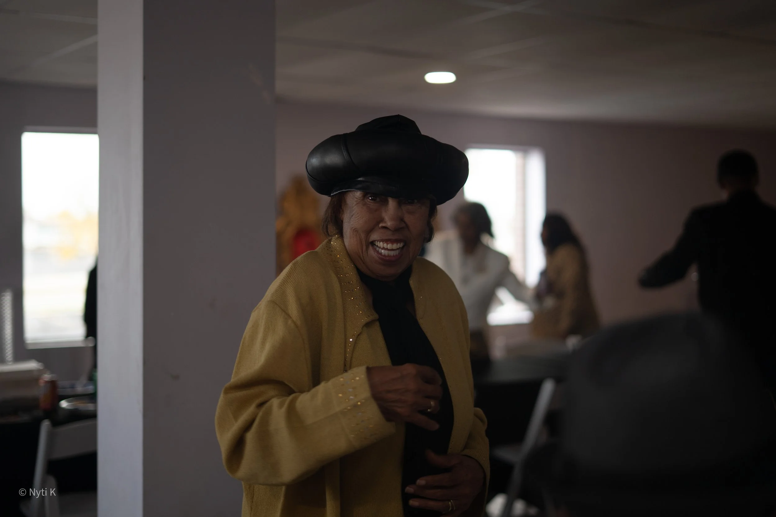 A smiling woman wearing a black hat and a yellow jacket, standing indoors with other people in the background.