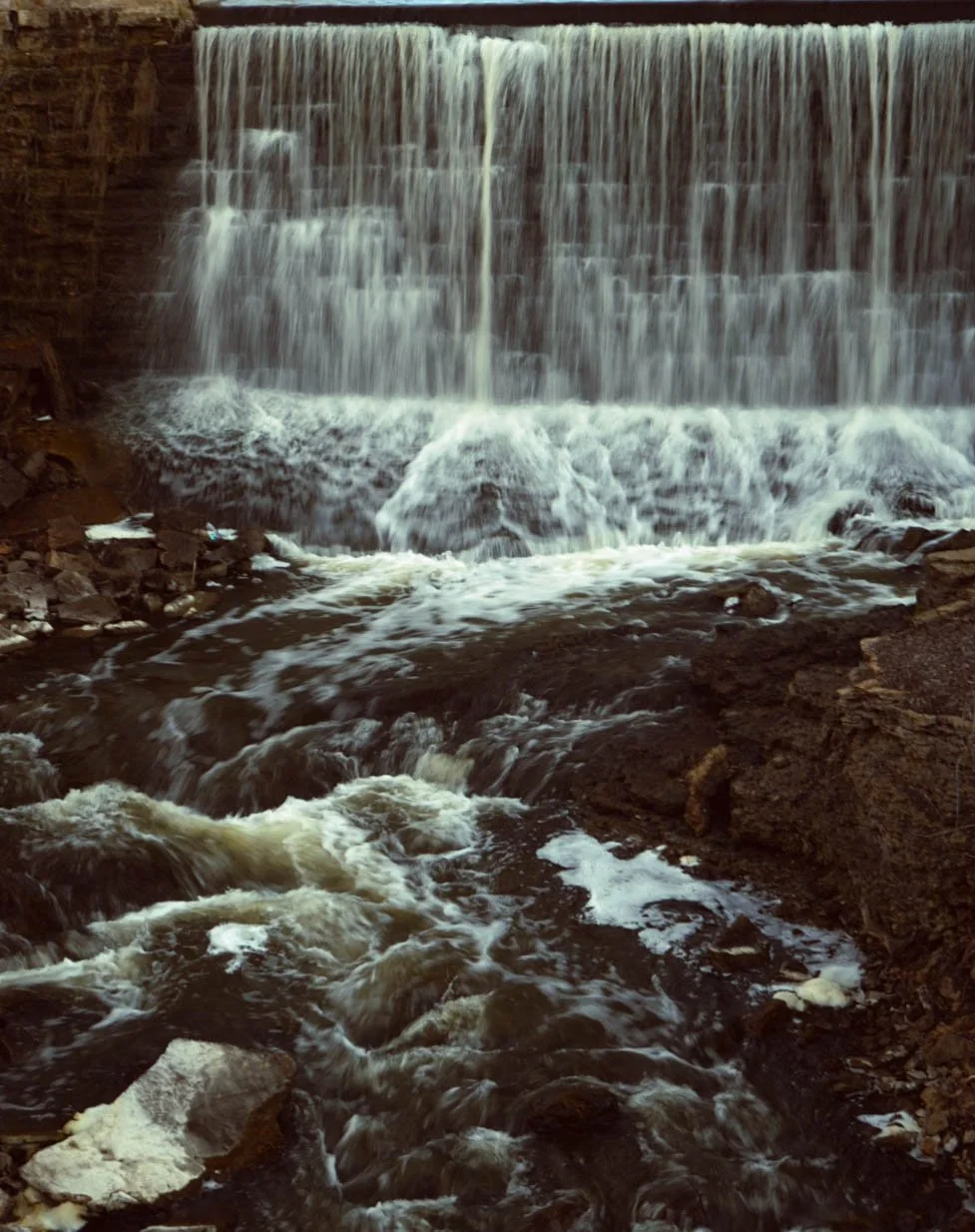 Waterfall flowing over a rocky ledge into a turbulent river below.