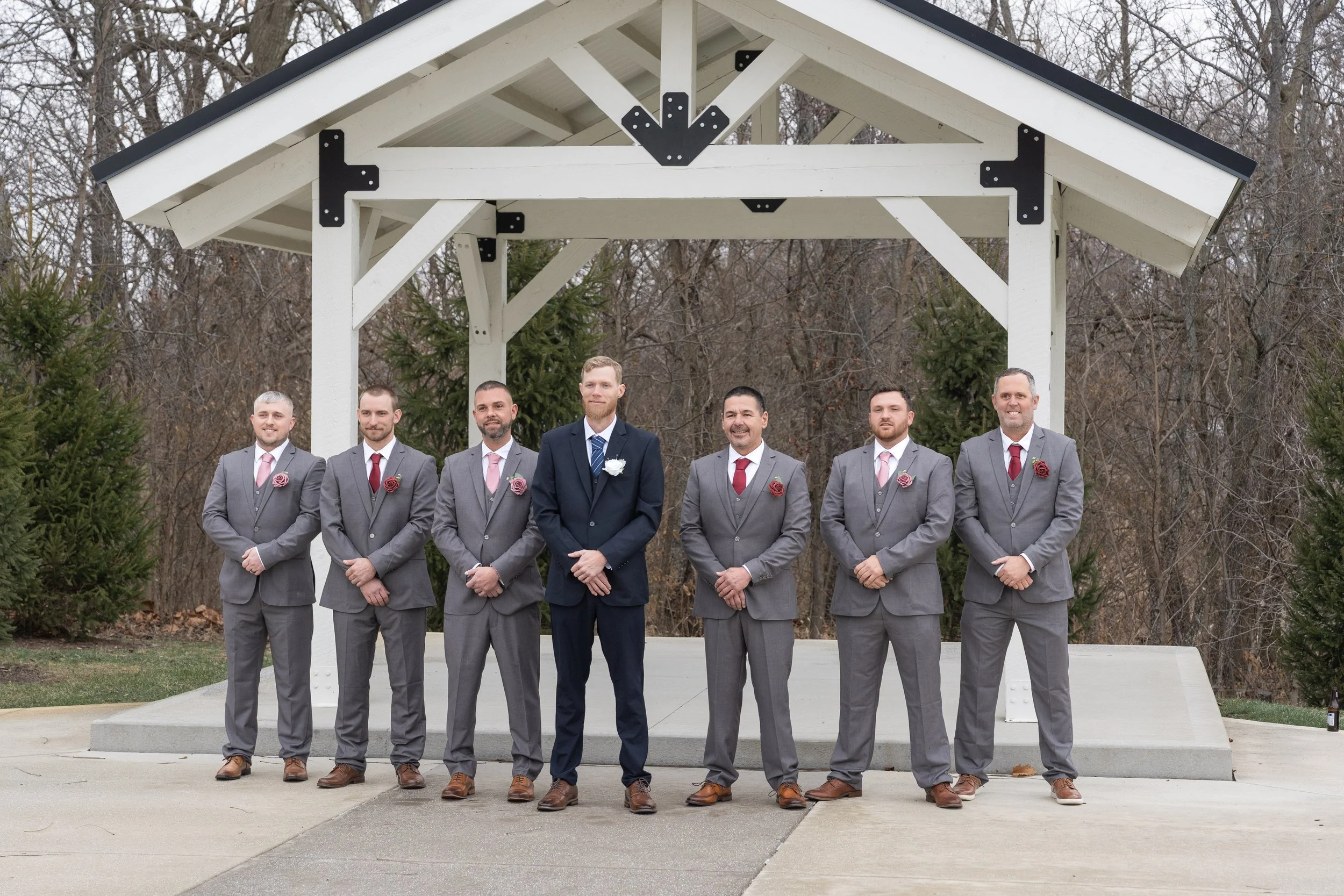 A group of seven men dressed in gray suits, with one man in a navy blue suit, standing outdoors under a white pavilion with a peaked roof, surrounded by bare trees and some greenery, during what appears to be a formal or wedding event.