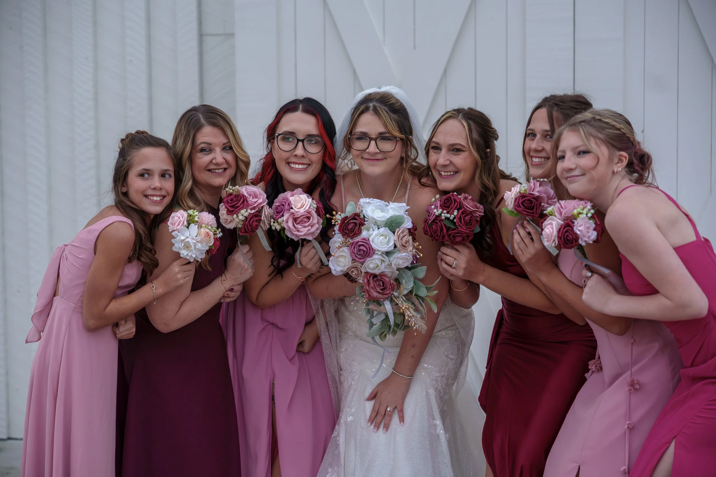 A bride surrounded by seven women, all holding bouquets of pink, purple, and white roses, standing in front of a white wall with geometric patterns.