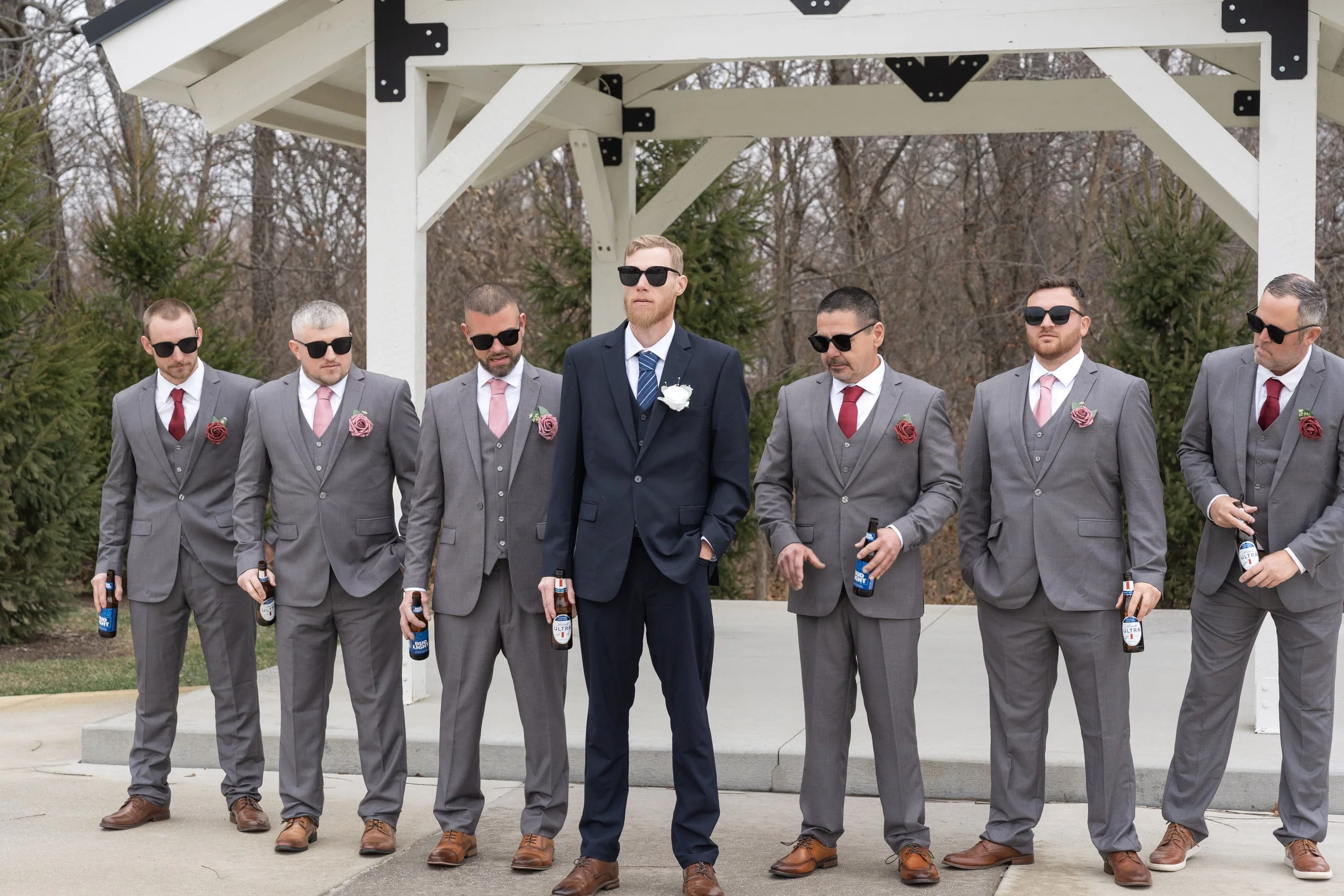A group of seven groomsmen wearing gray suits, pink and red boutonnieres, and black sunglasses, standing outdoors in front of a white gazebo structure. The groom, in a navy suit with a white boutonniere, is at the center.
