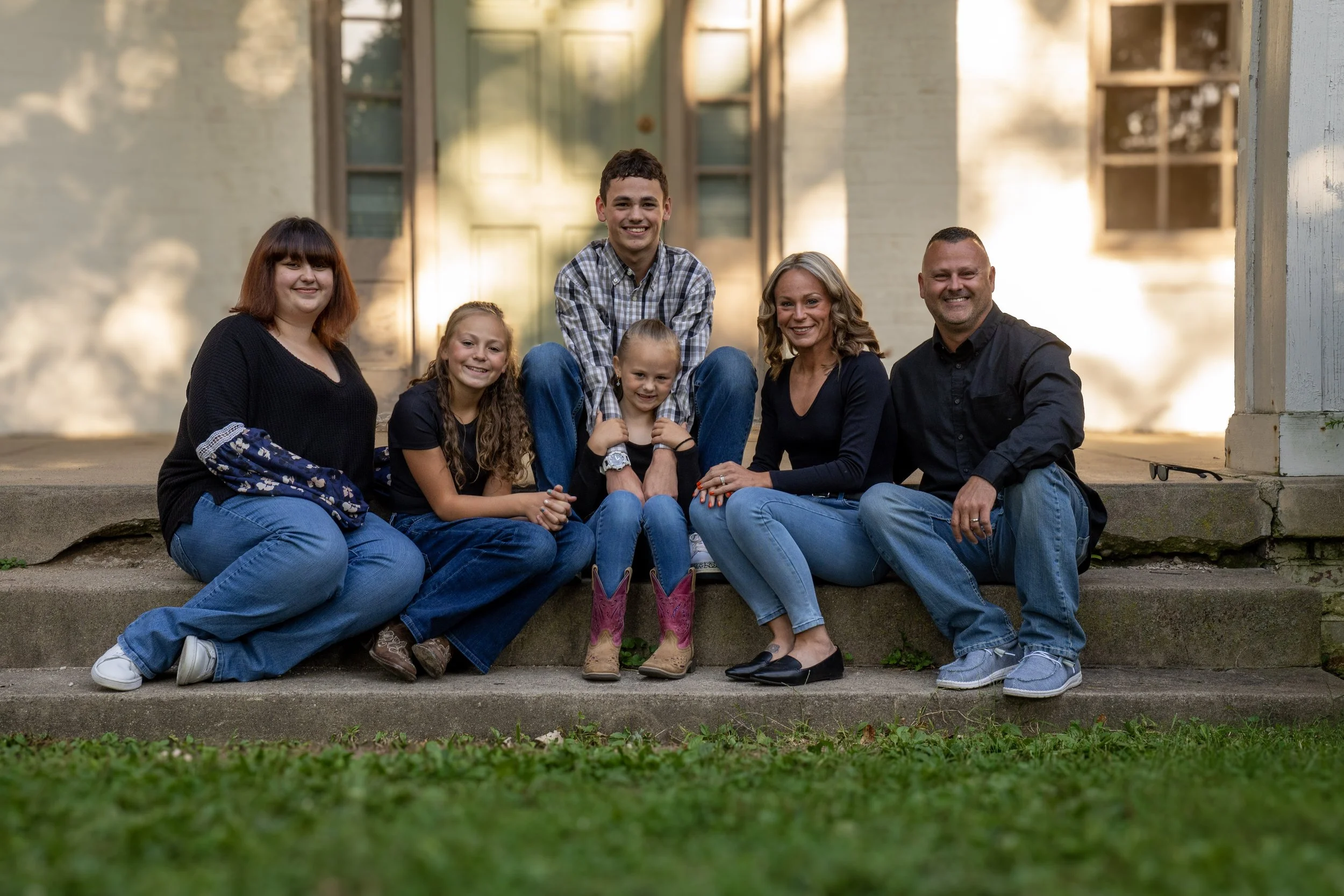 A family of seven sitting on the steps of a porch in front of a house with a yellow door. The family includes three adults and four children, all smiling and looking at the camera. The setting is in the late afternoon or early evening with warm lighting.