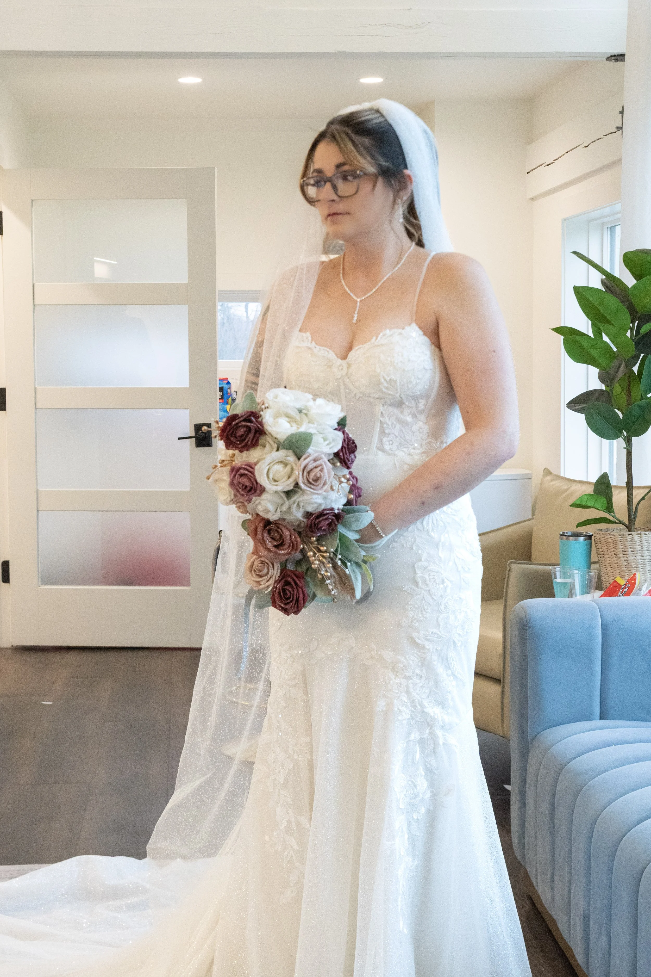 A bride wearing glasses, a veil, and a white wedding dress with lace details is holding a bouquet of white and dark pink roses. She is standing indoors near a white sliding door, with a green potted plant and a blue chair visible in the background.