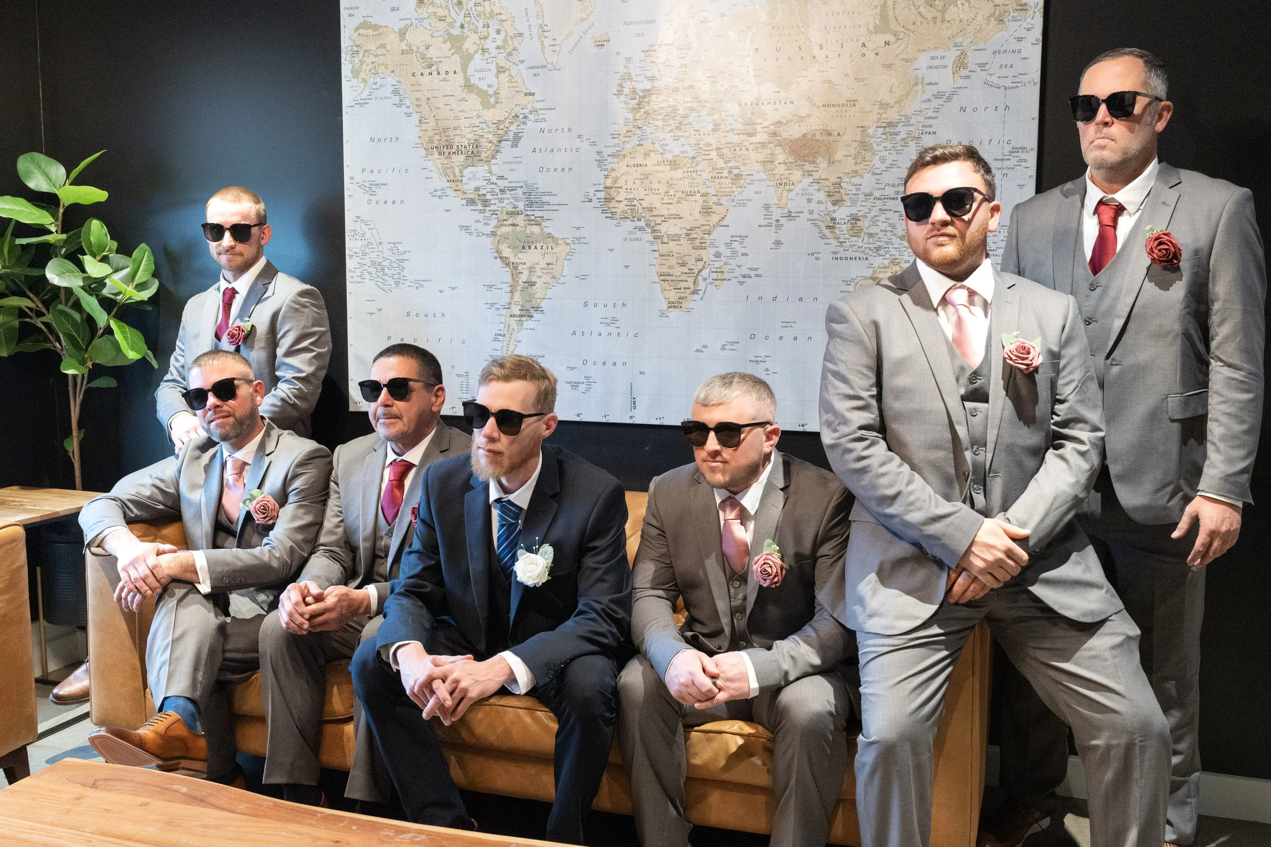 Seven men in suits with pink or red roses on their lapels, wearing sunglasses, are gathered in front of a world map. Four men are seated, and three are standing.