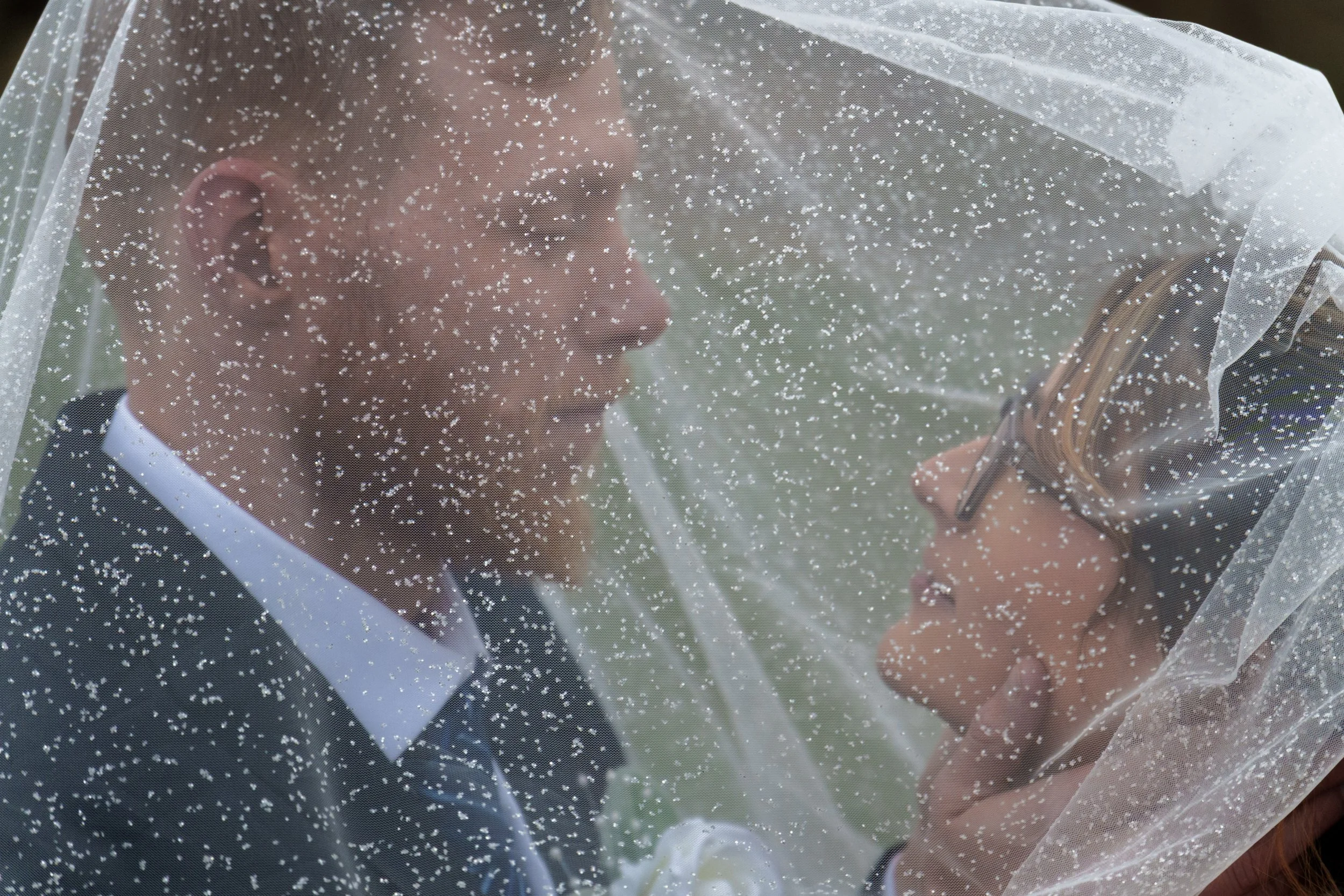 A bride and groom dressed in wedding attire, with the bride holding a bouquet, under a veil with white dots, during their wedding.