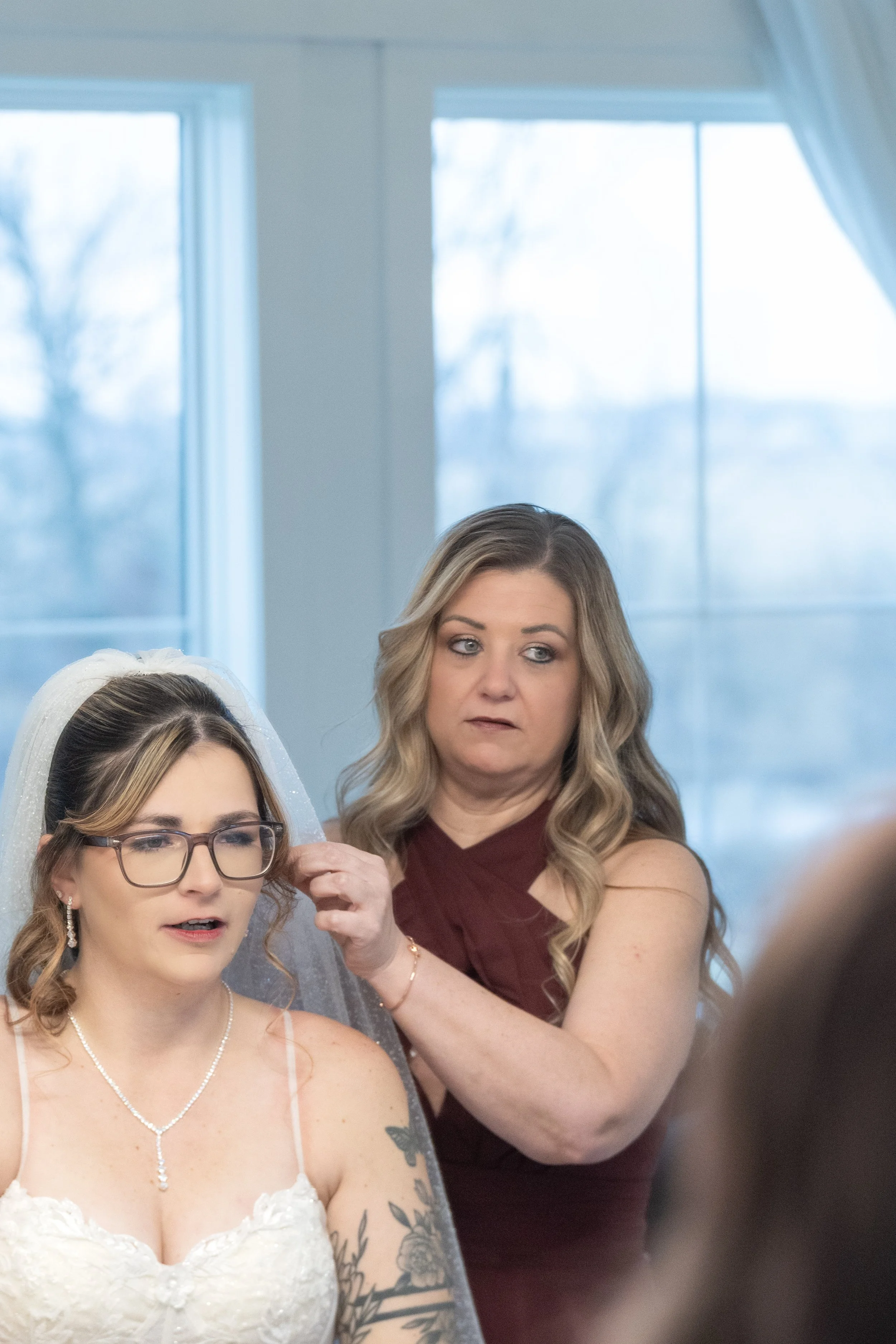 A woman in a wedding dress getting her hair done by another woman in a sleeveless maroon dress, with scenic window view in the background.