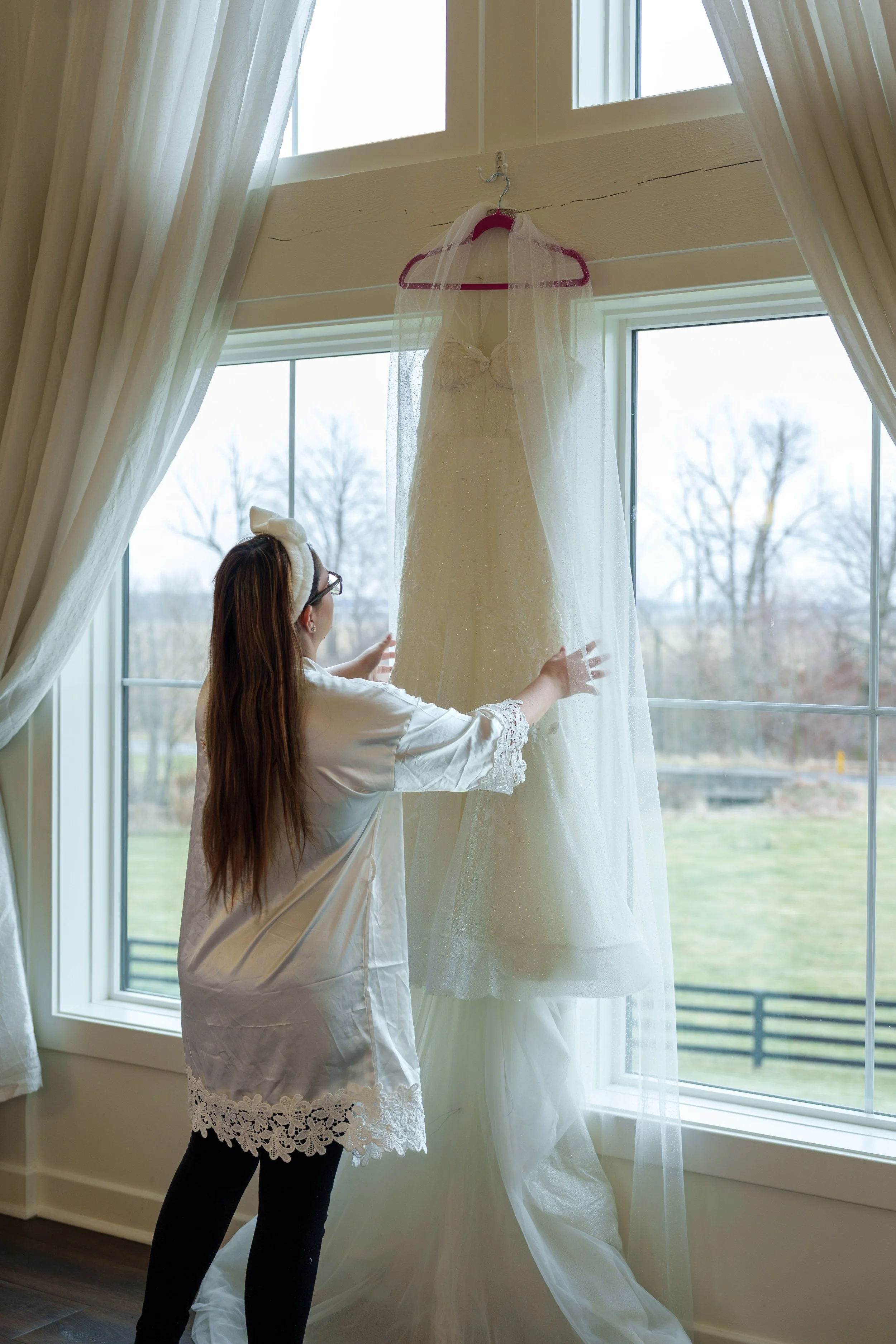 A woman with long hair, glasses, and a headband is standing by a window, adjusting a wedding dress hanging on the curtain rod. The wedding dress is ivory or white, with lace details, and a sheer veil.