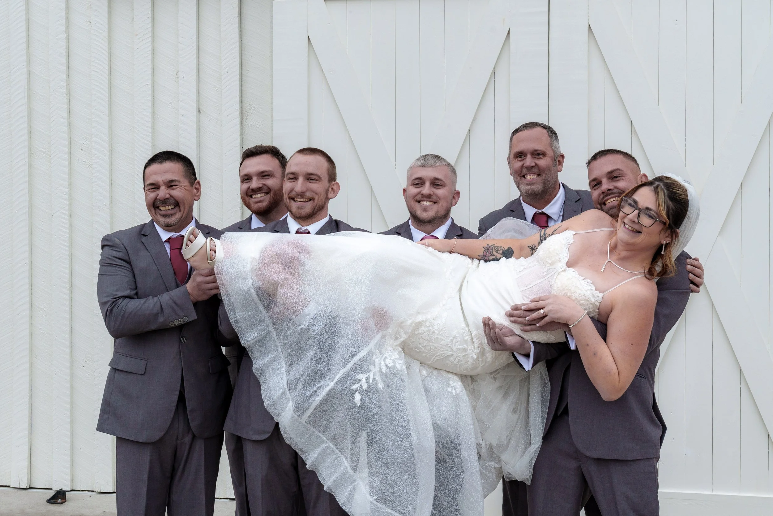 A group of six men in suits holding a smiling woman in a wedding dress in front of a white wooden wall.
