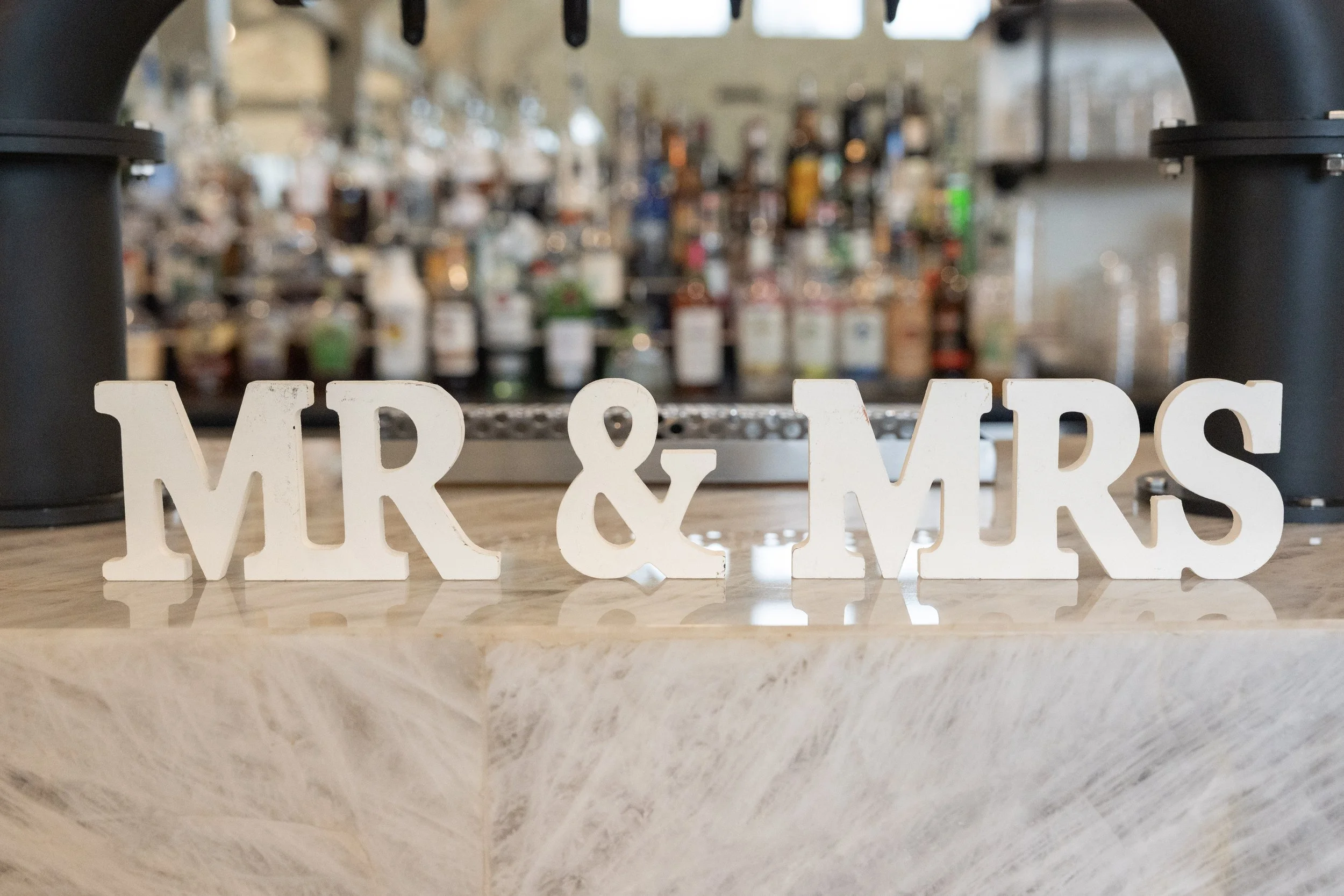 White decorative letters spell out 'MR & MRS' on a marble surface, with a blurred background of a bar or kitchen area with various bottles.