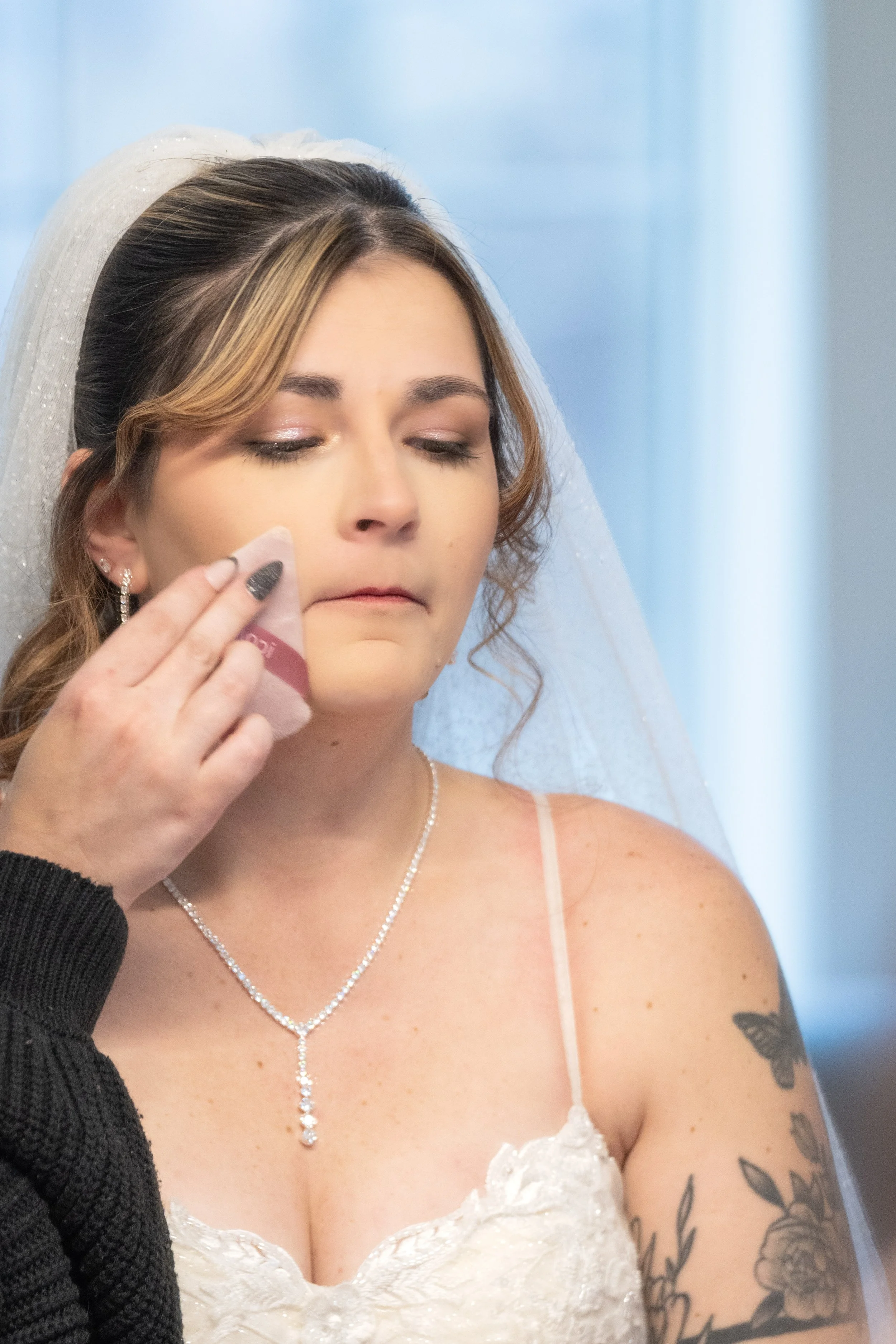 A bride with closed eyes, wearing a white lace wedding dress and jewelry, receives makeup or skincare treatment.
