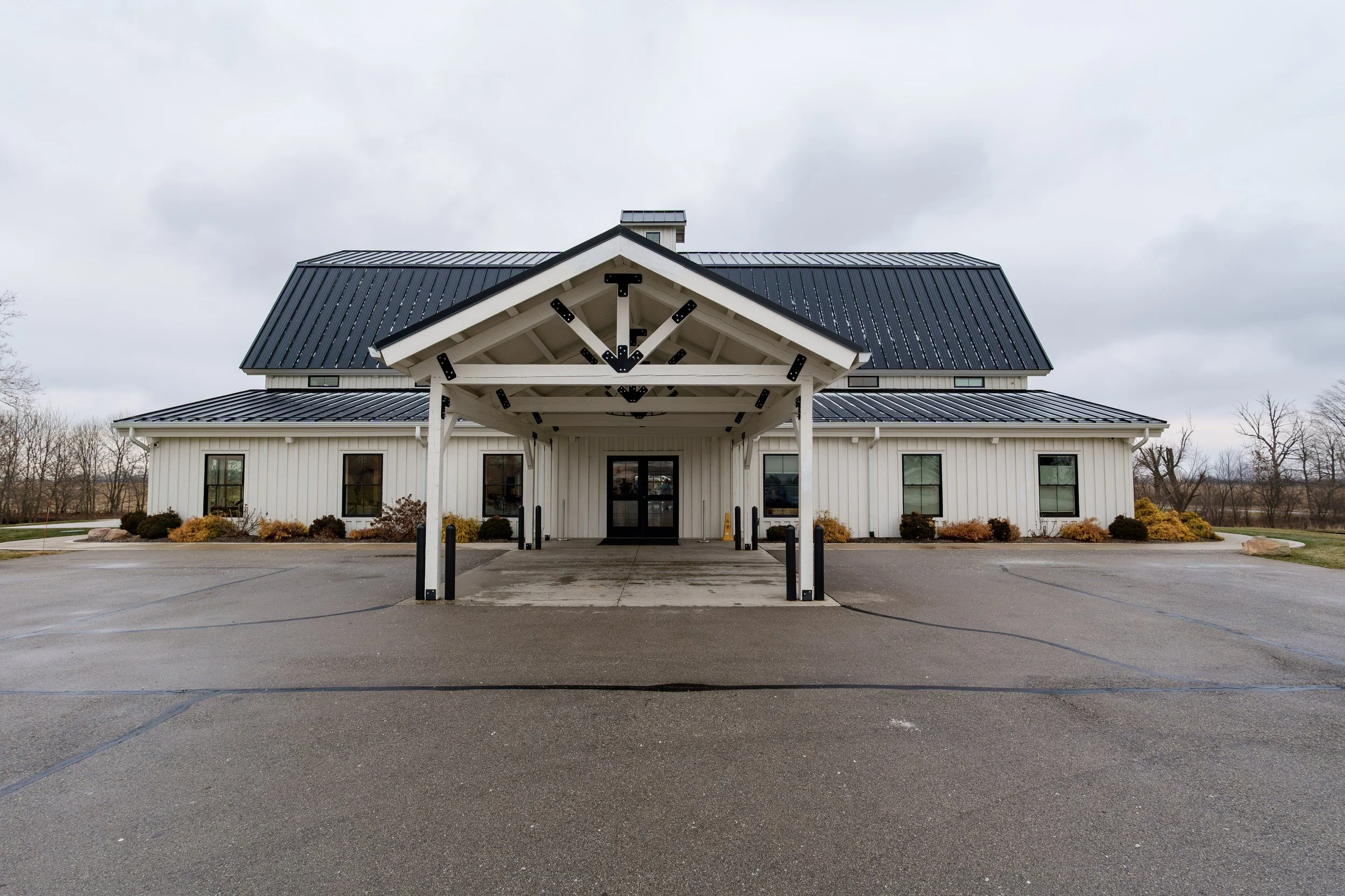 Front view of a white building with a covered entrance, black metal roof, and surrounding landscaped bushes, under a cloudy sky.