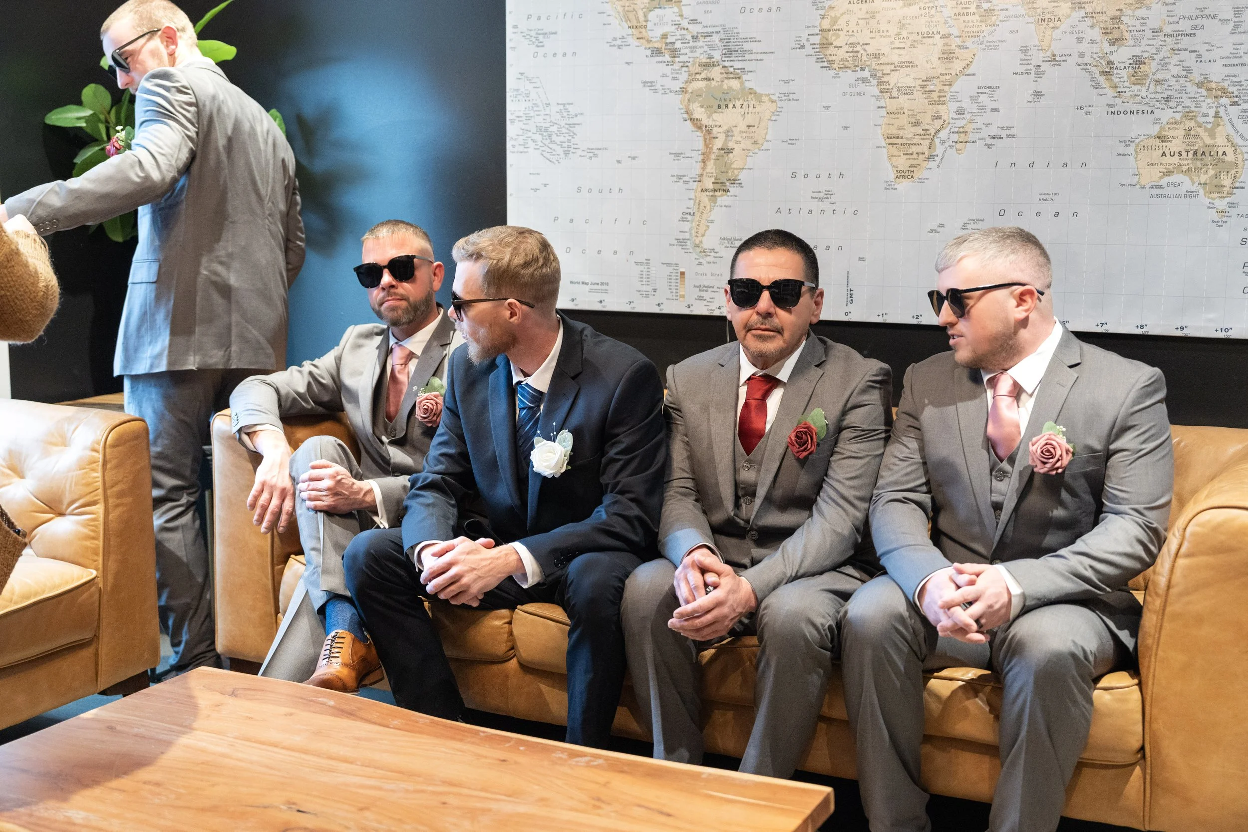 Five men dressed in suits sitting on a tan leather couch in a room with a world map on the wall, wearing sunglasses and flower boutonnieres, likely at a formal event.
