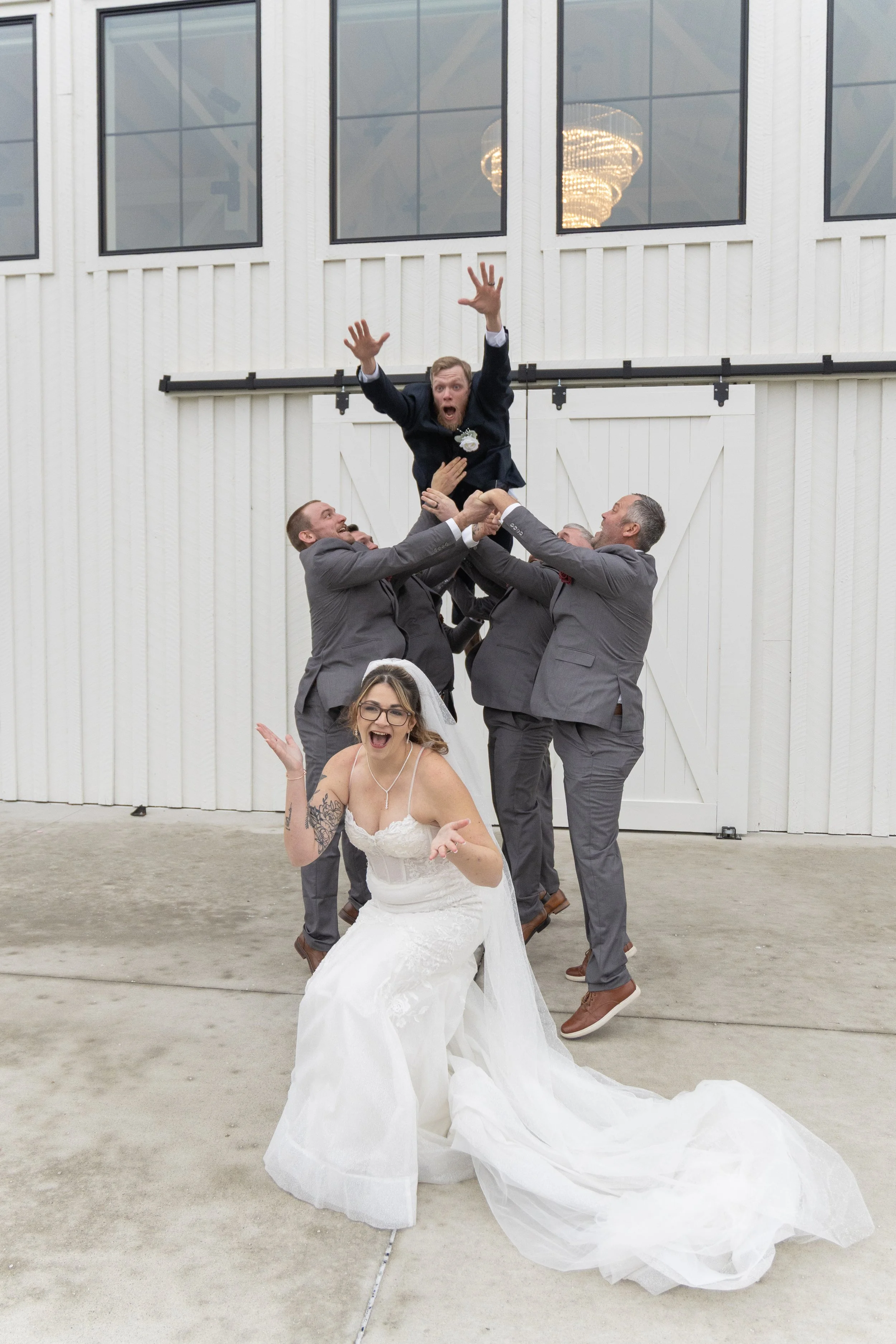 A bride in a white wedding dress with tattoos on her arms, performing a joyful dance in front of a group of men in suits who are lifting a man in a suit above their heads outside a white barn. The man being lifted looks surprised and excited.