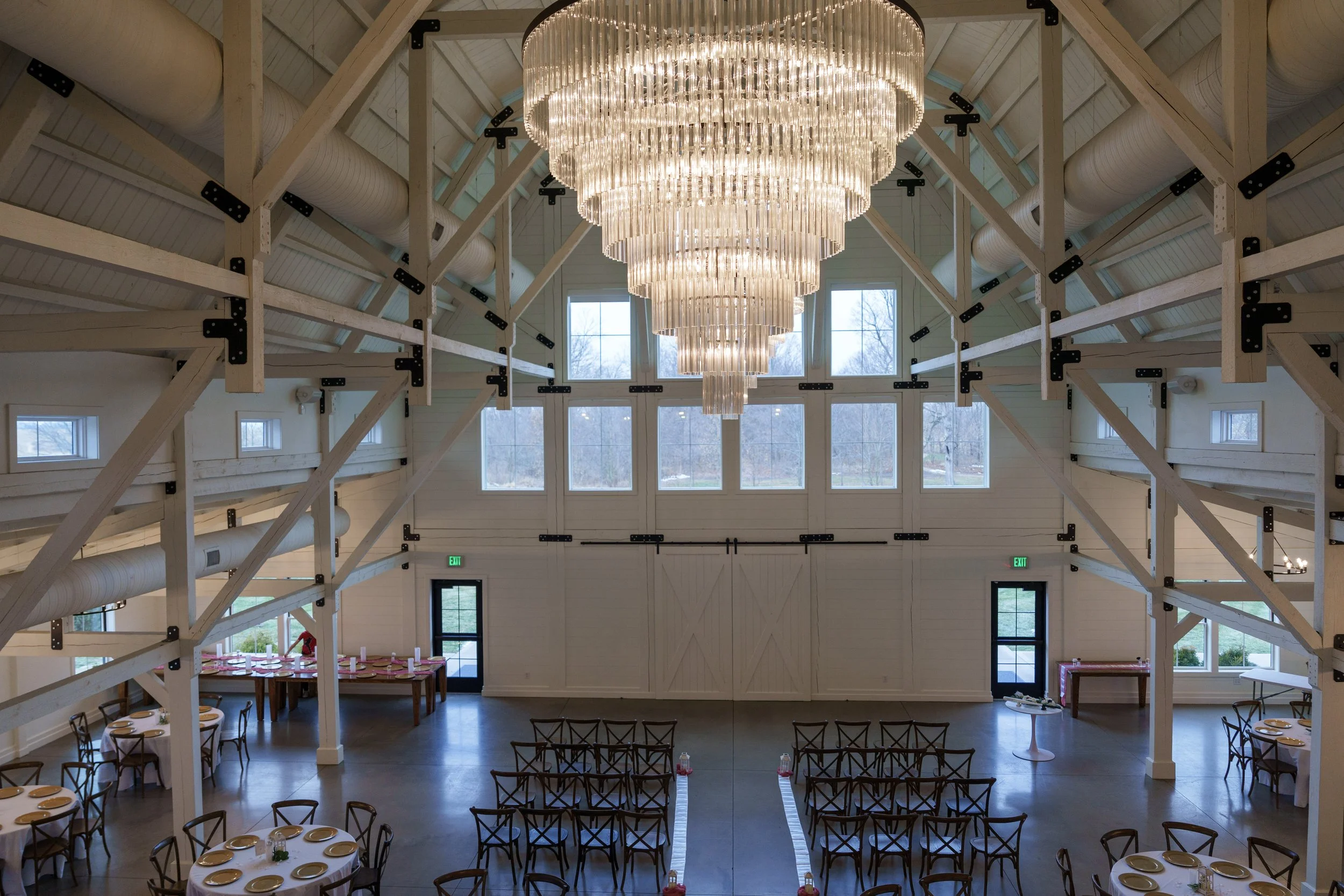 Interior view of a spacious event hall with a large chandelier hanging from the ceiling, featuring multiple tiers of lights. The hall has high ceilings with exposed beams and large windows letting in natural light. There are decorated tables set with