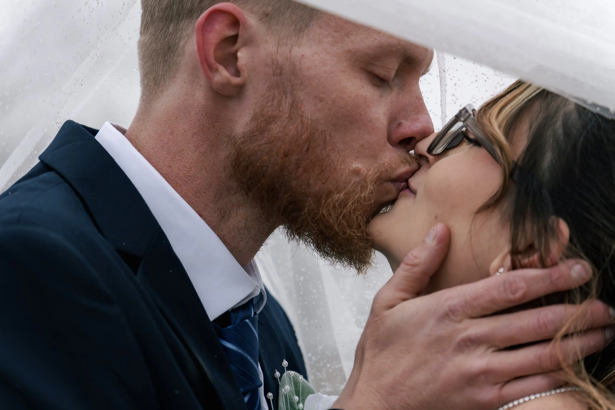 A man and a woman are sharing a kiss at a wedding, with their eyes closed and smiling. The man has a beard and is wearing a dark suit with a white shirt, while the woman has glasses and long hair, and is wearing wedding jewelry.