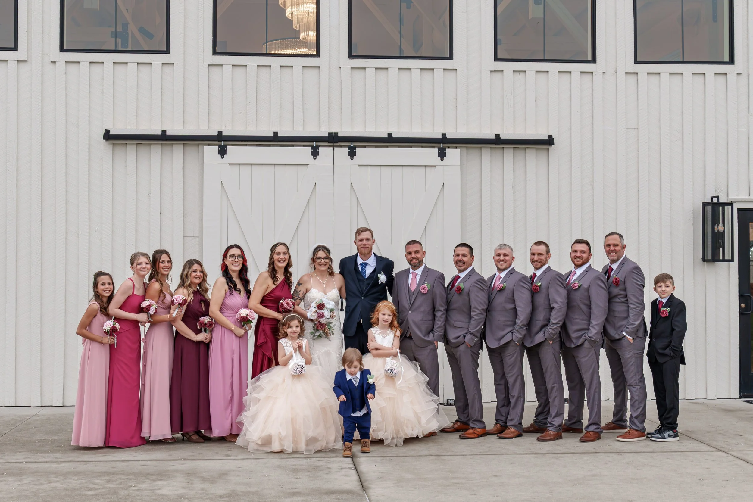 A large group of people, including adults, children, and a bride and groom, gathered in front of a white barn facade during a wedding celebration.