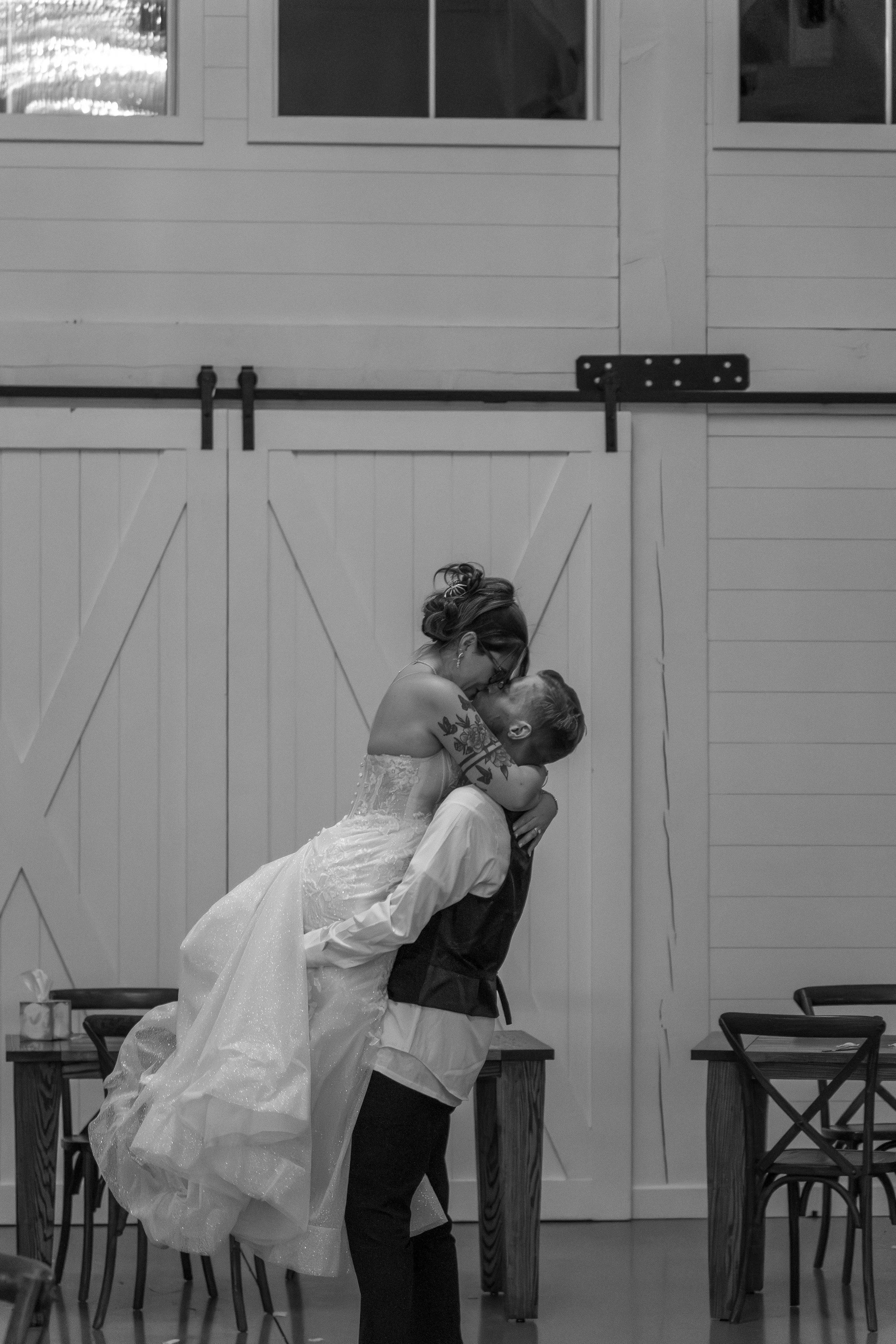 A couple, where the woman is dressed in a wedding gown and the man in dress clothes, is sharing a kiss inside a rustic, barn-like venue with wooden paneled walls and tables, and sliding barn doors in the background.