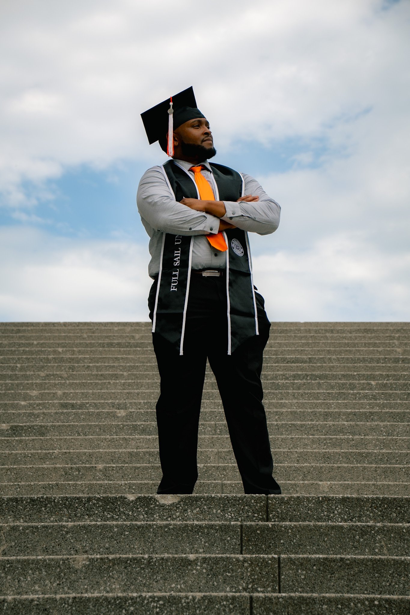 A young man in a graduation cap and gown standing on outdoor stairs with arms crossed, looking proud and confident against a cloudy sky.