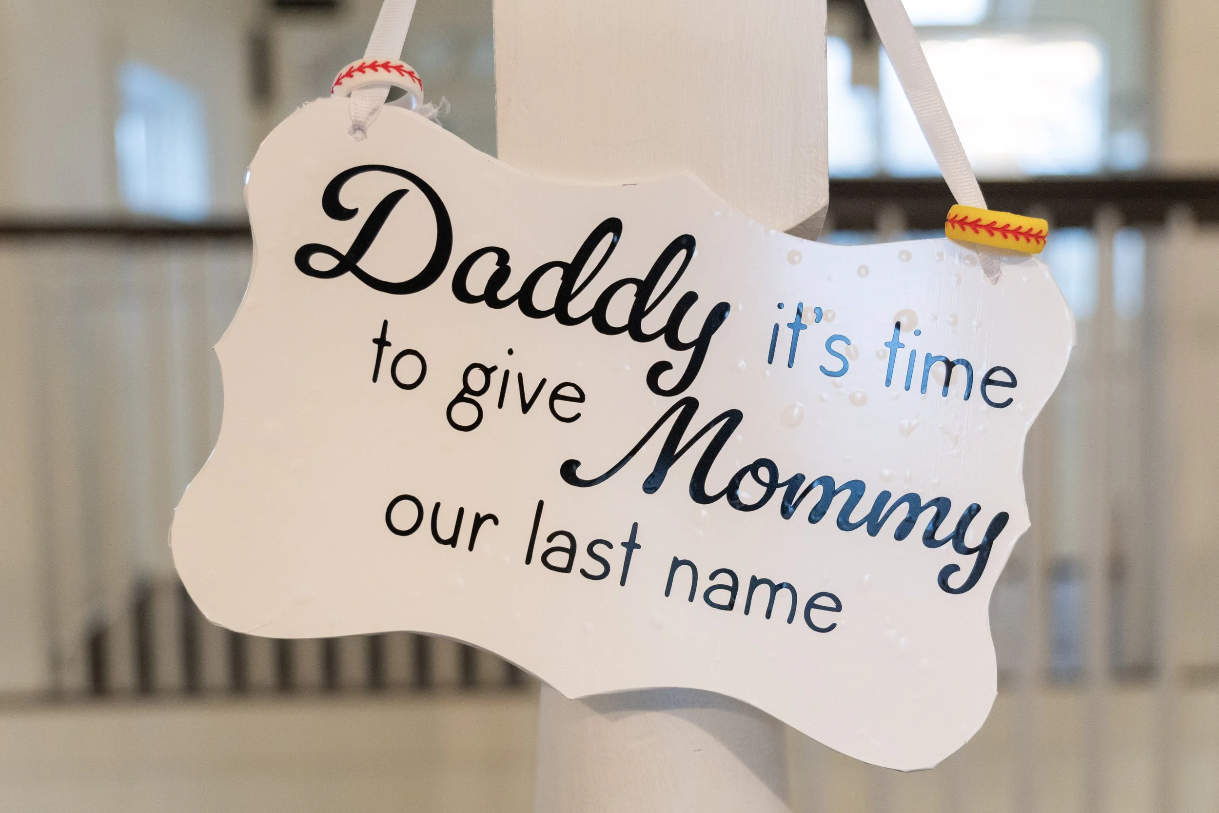 A decorative sign shaped like a cloud with the words 'Daddy, it's time to give Momma our last name' written on it, hanging from white ribbon with red and yellow baseball-themed accents.