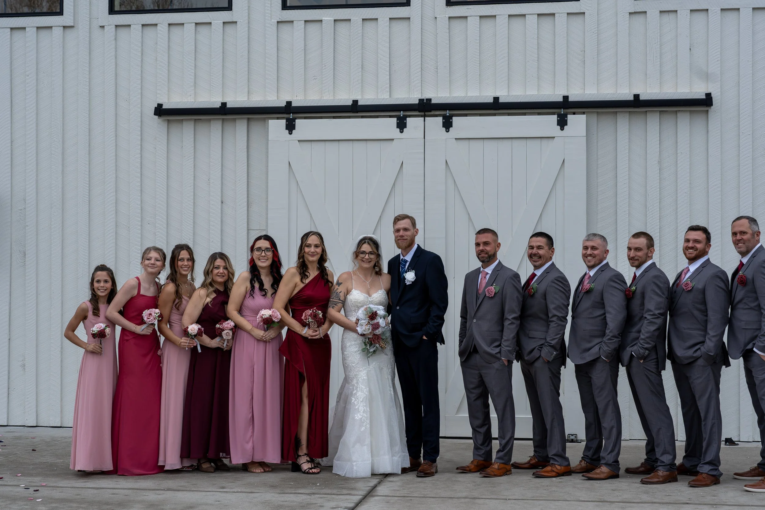 A group of people dressed for a wedding standing in front of a white wooden barn door, with women in pink and red dresses and men in gray suits and ties.