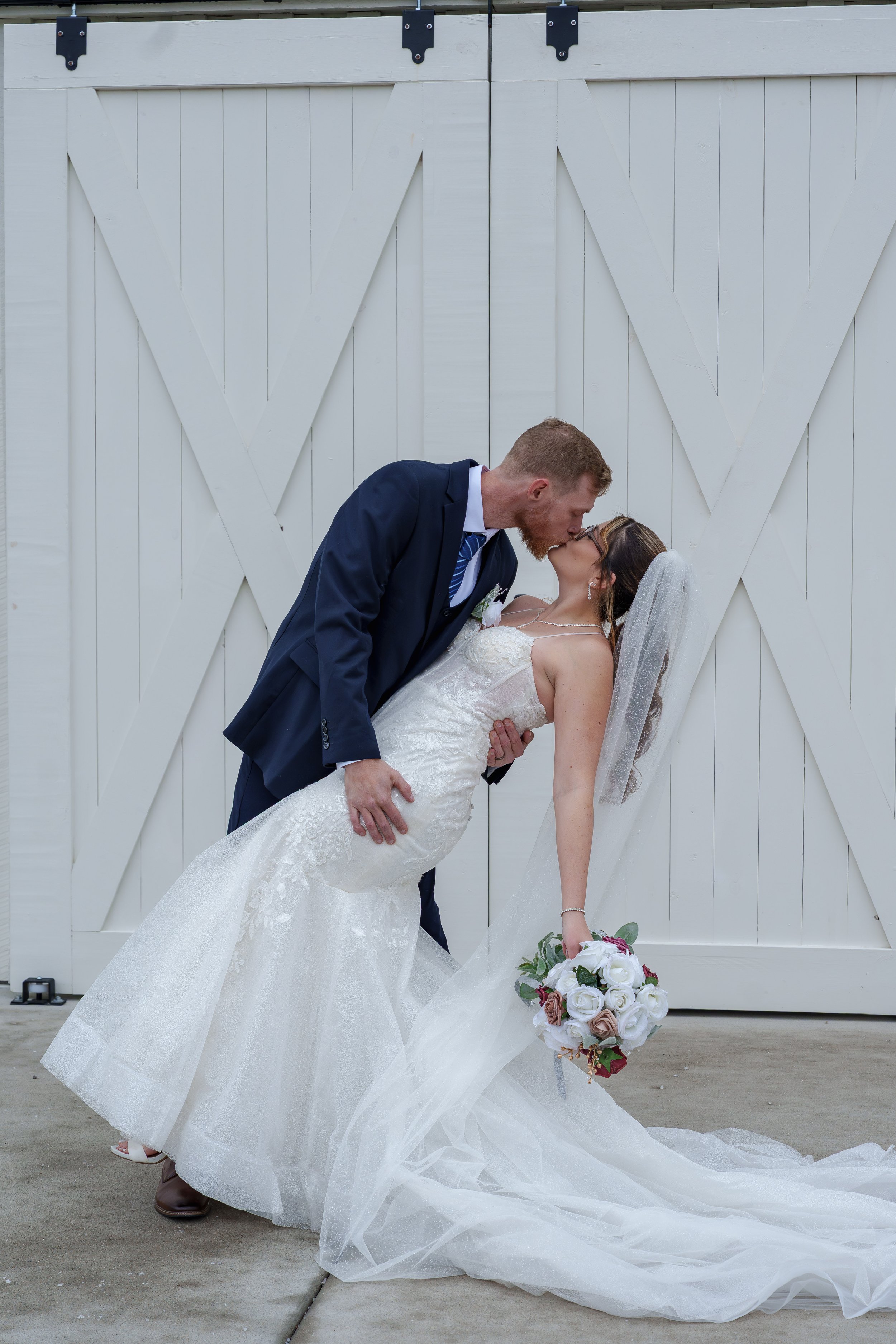 A newlywed couple shares a kiss in front of a white barn door. The bride wears a white wedding dress with lace details, a veil, and holds a bouquet of white and blush roses. The groom is dressed in a navy suit with a tie and boutonniere. The man is l