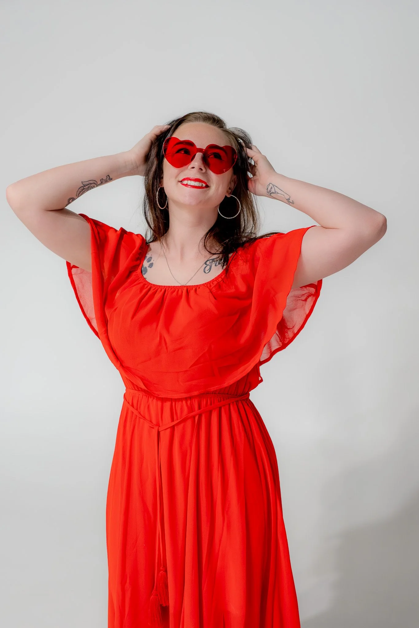 A woman wearing a bright red dress and oversized red sunglasses, smiling and holding her hair back, against a plain light grey background.