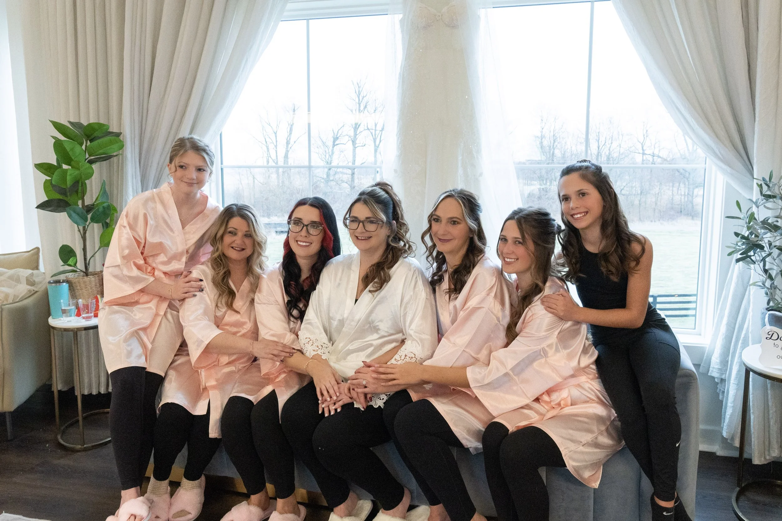 A group of seven women, some wearing pink satin robes, gather in a bright room with large windows and white curtains. They are smiling and posing together for a photo.