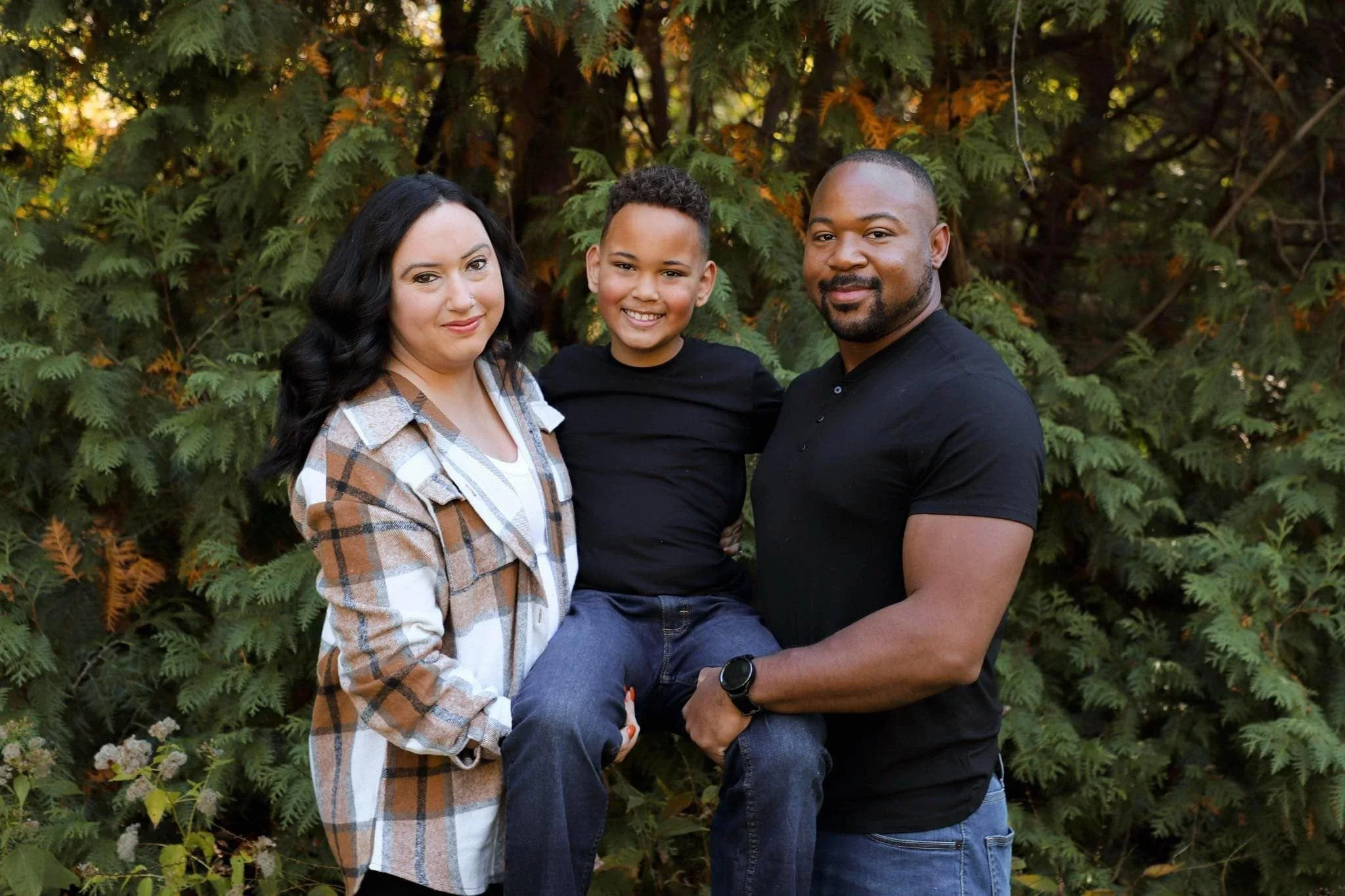 A smiling family of three, including a woman with dark hair and a man with a beard holding a young boy with curly hair, standing outdoors in front of green foliage.