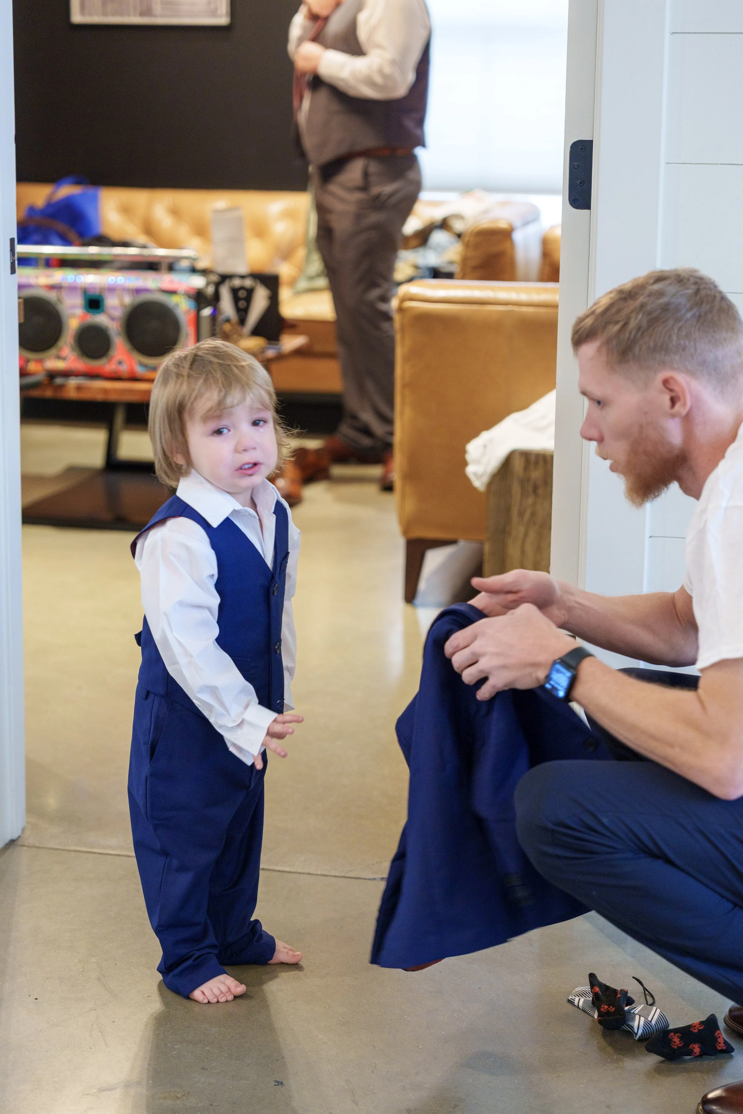 A young boy with blonde hair and a white shirt under a blue vest, standing barefoot on a beige floor, looking at a man who is sitting and folding a dark blue suit jacket. In the background, there is a woman with a handbag, and various objects includi