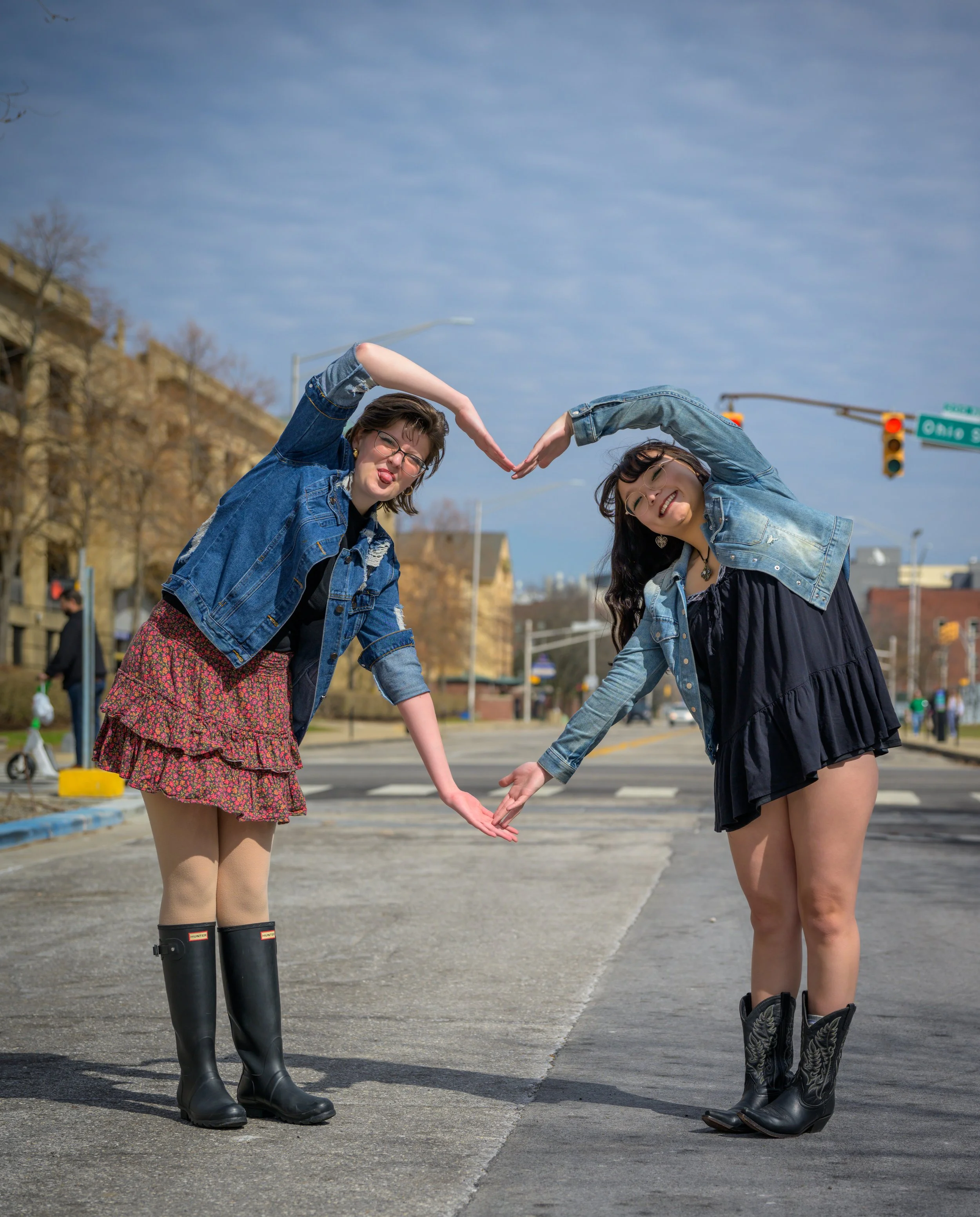 Two women standing on a street, bending down and touching hands to form a heart shape, smiling, wearing denim jackets and boots, with traffic lights and buildings in the background during daytime.