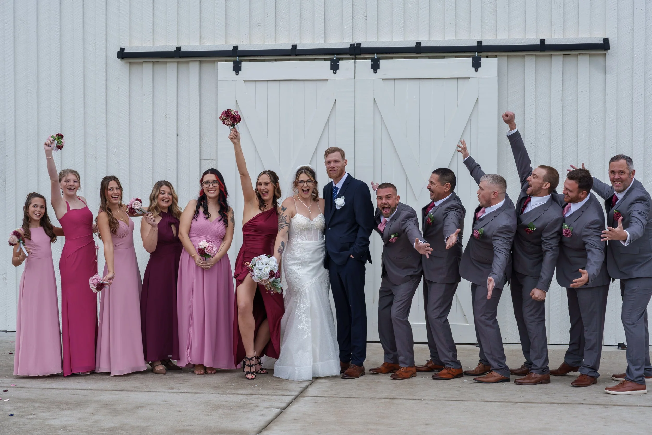 A group of happy wedding guests, including bridesmaids in pink dresses and groomsmen in gray suits, celebrating with the bride and groom in front of a white barn door.