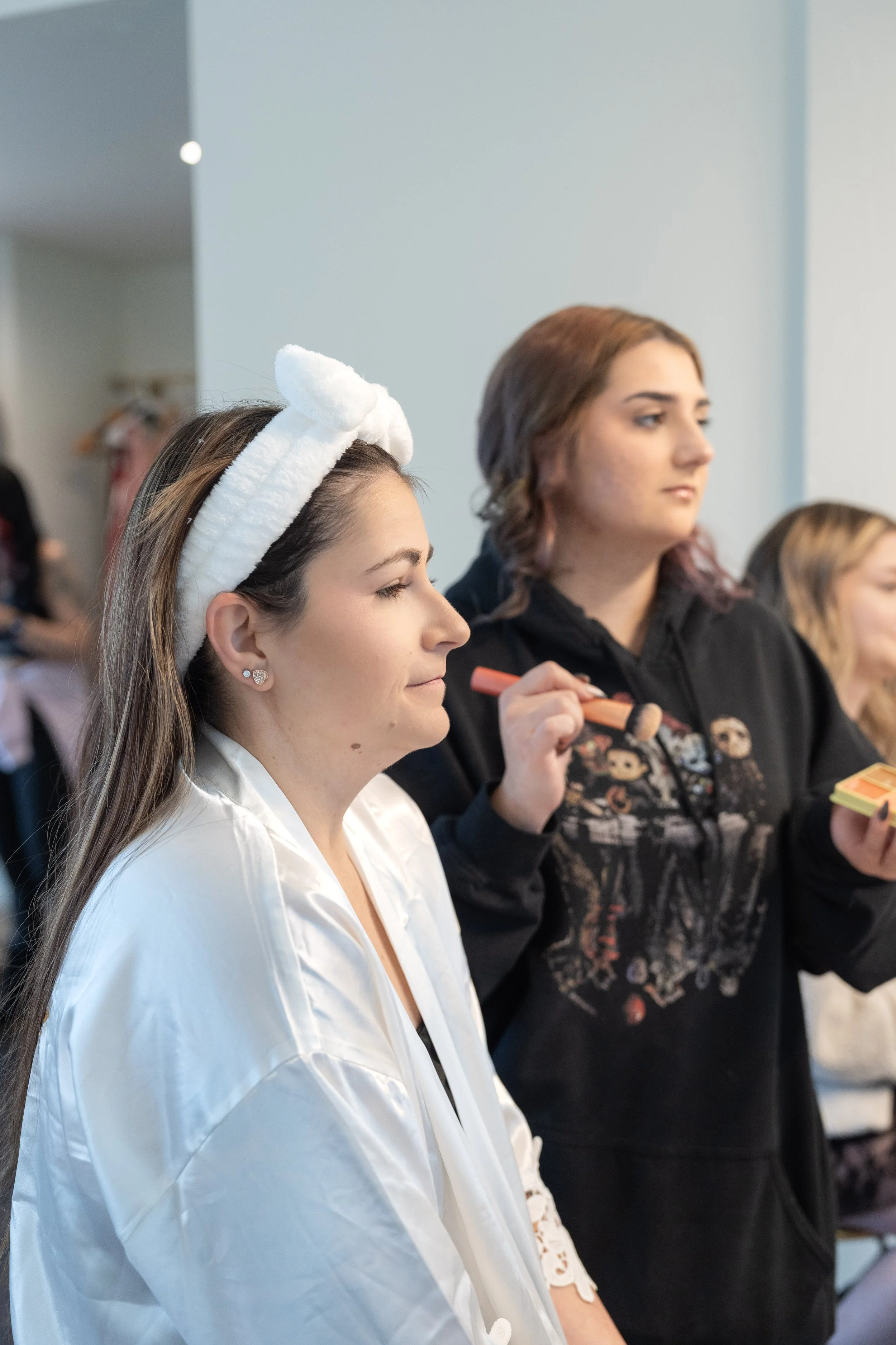 Two women sitting side by side, one with a white fluffy headband and the other with a black hoodie, possibly in a beauty or makeup setting.