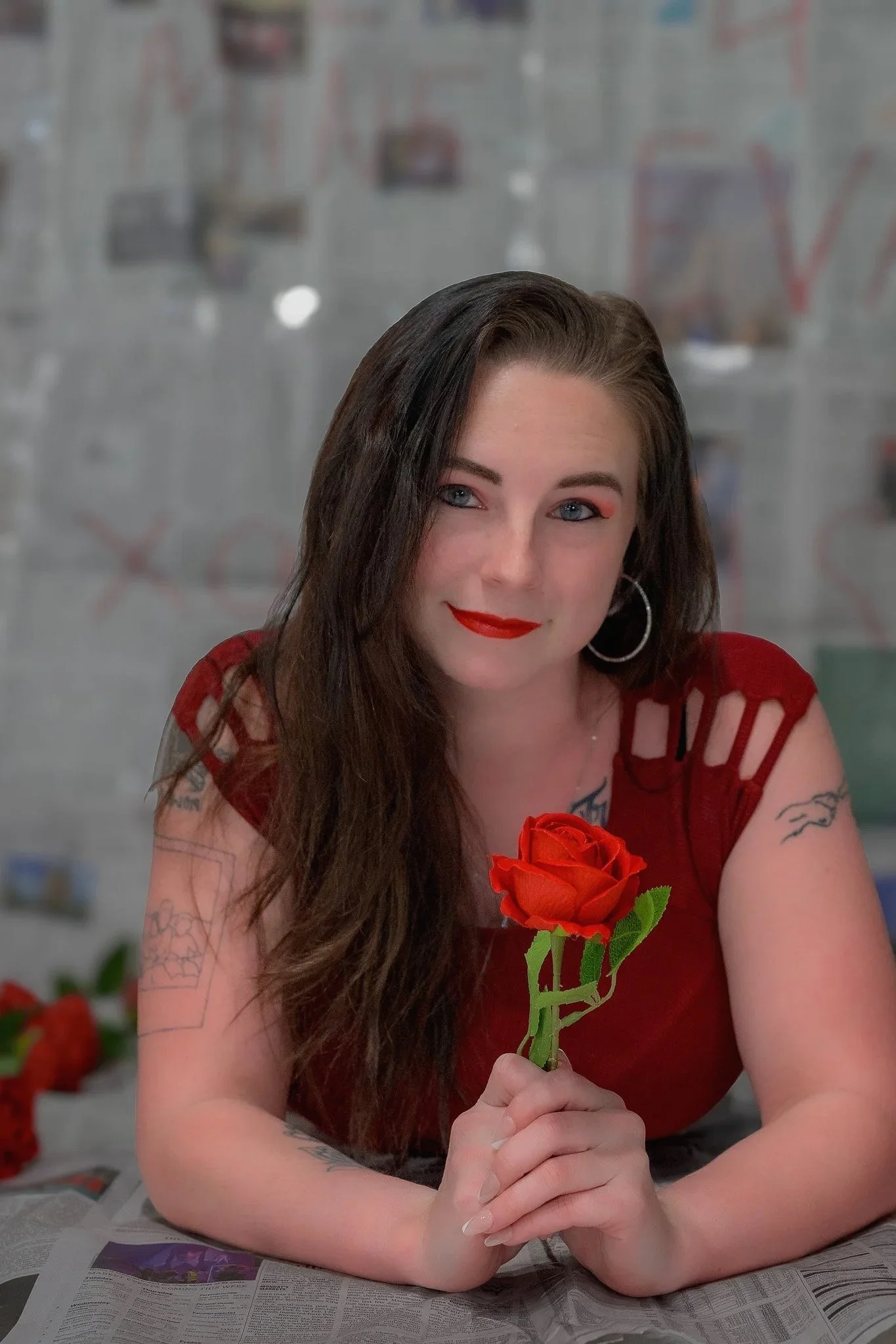 A woman with long brown hair, red lipstick, and hoop earrings, sitting at a table covered with newspapers, holding a red rose in her hands. She is wearing a red top with cut-out shoulders.