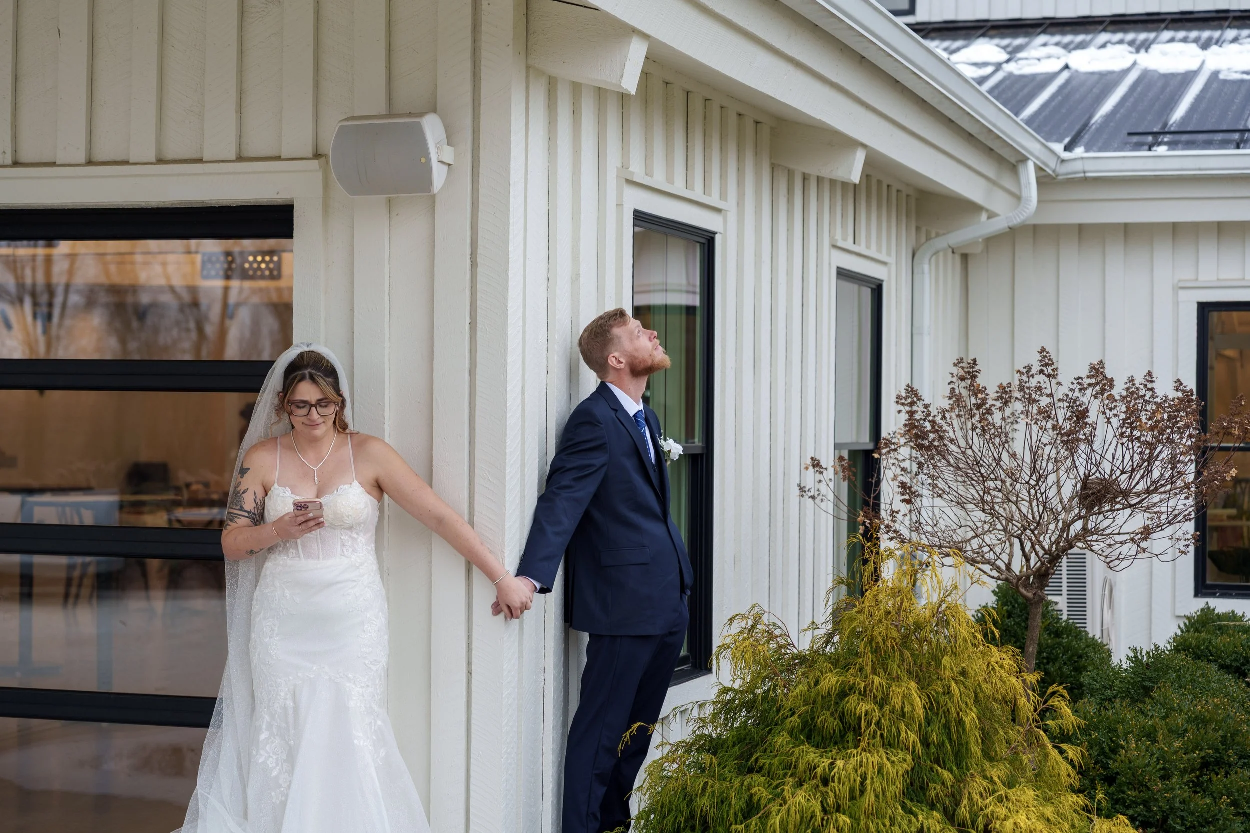 A bride and groom stand side by side outside a house, holding hands. The bride is wearing a white wedding dress with rosé hair, glasses, and has tattoos on her arms. The groom is dressed in a dark blue suit, white shirt, blue tie, and a white flower 