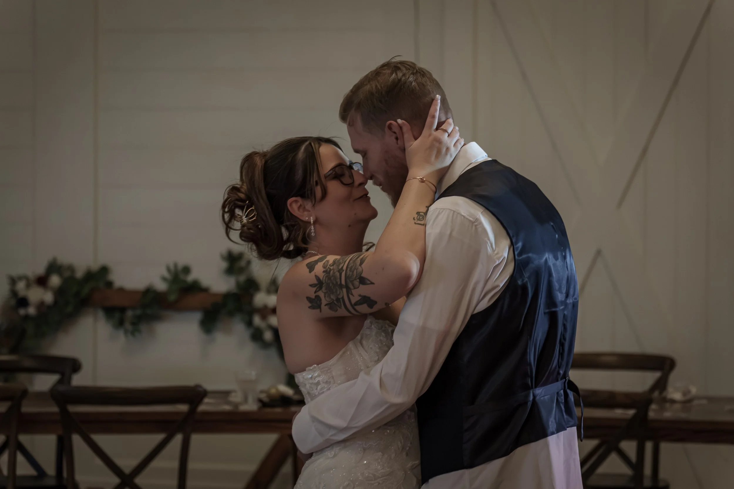 A bride and groom sharing a dance, close and intimate, in a wedding reception setting with decorated tables and chairs in the background.
