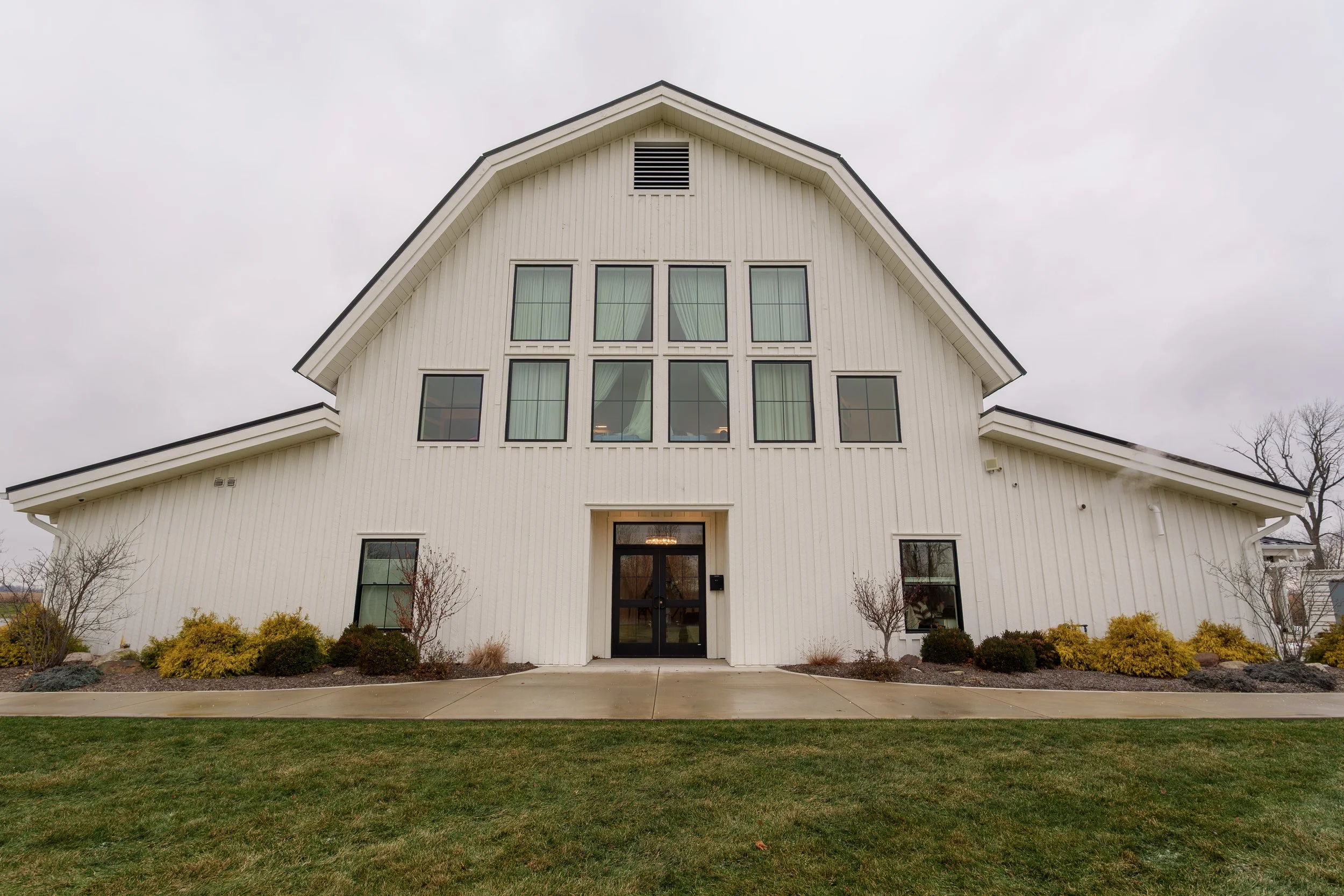 Front view of a large white modern house with multiple windows, a black door, and landscaped bushes in the front yard, under an overcast sky.