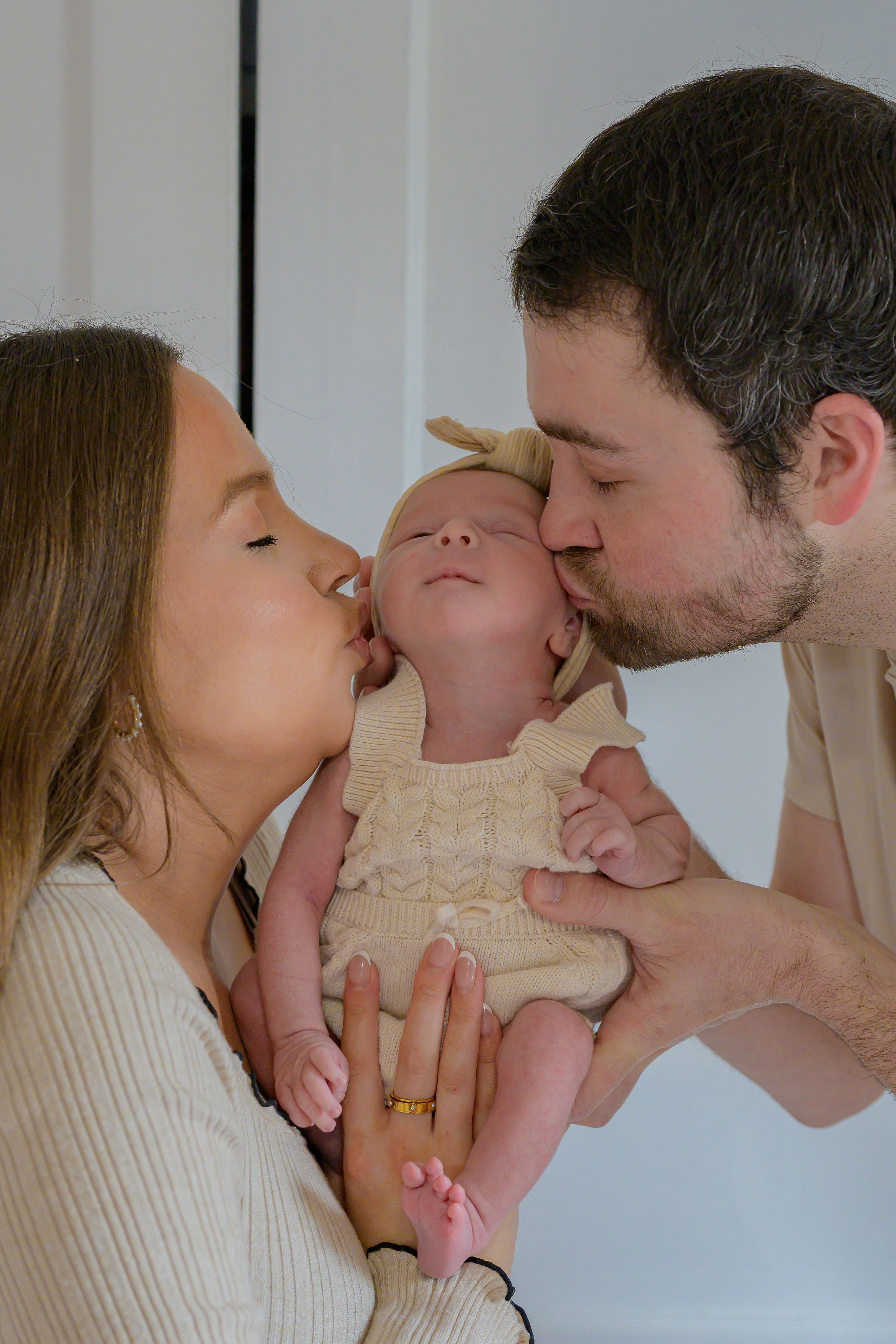 A family holding a newborn baby together, with the mother on the left, the father on the right, and the baby in the middle, all touching foreheads and lips touching the baby's face.