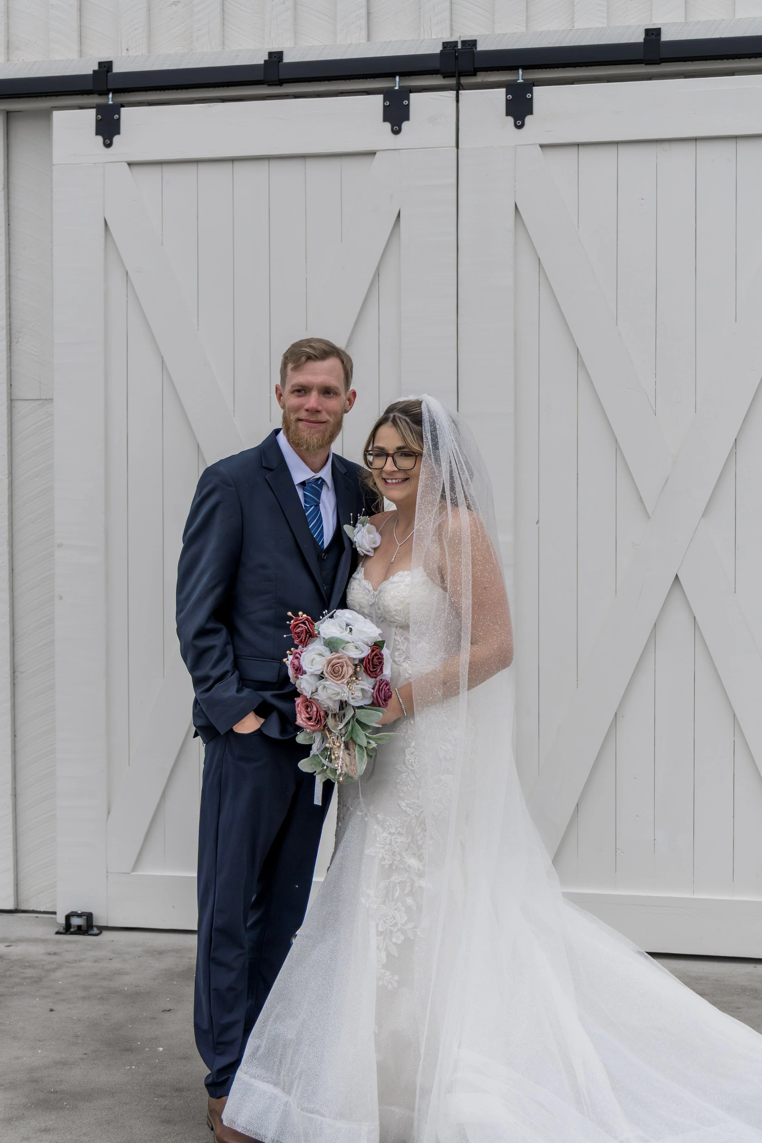 A bride and groom standing in front of a white barn door, dressed in wedding attire, smiling and holding a bouquet of flowers.