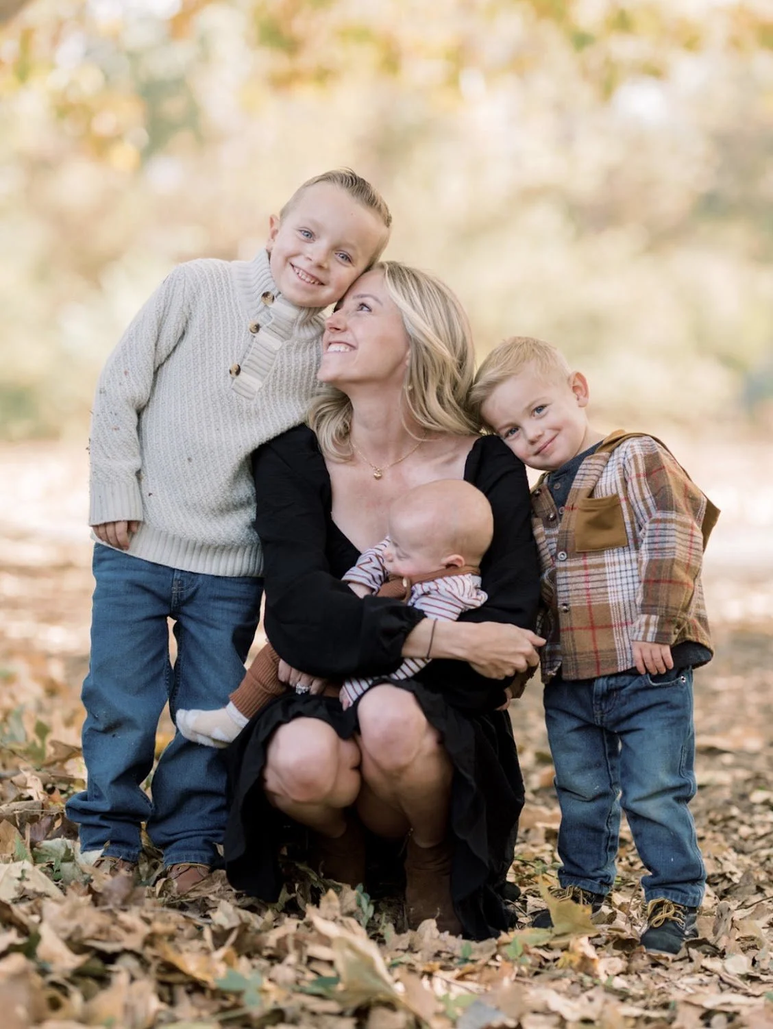 A woman with three young children outdoors in a park during autumn, surrounded by fallen leaves. The woman is holding a baby, smiling at two boys standing beside her. The children are dressed in fall clothing, and the background features blurred trees with colorful foliage.