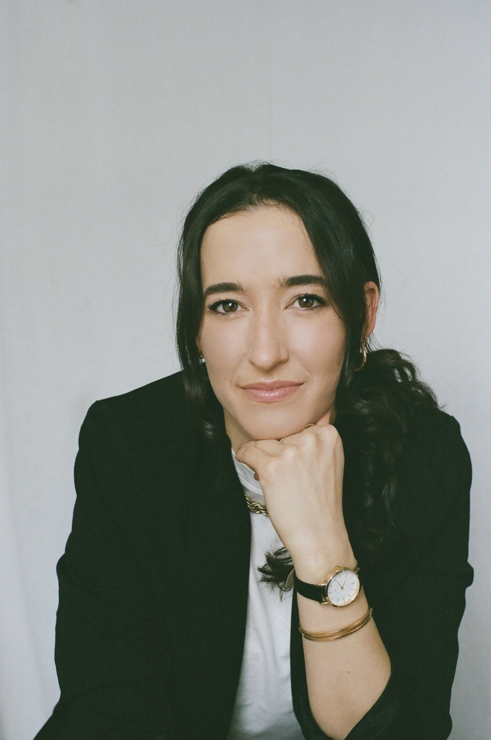 A woman with black hair, wearing a black blazer over a white top, resting her chin on her hand. She is wearing a gold watch and bracelets, and is in front of a plain white background.