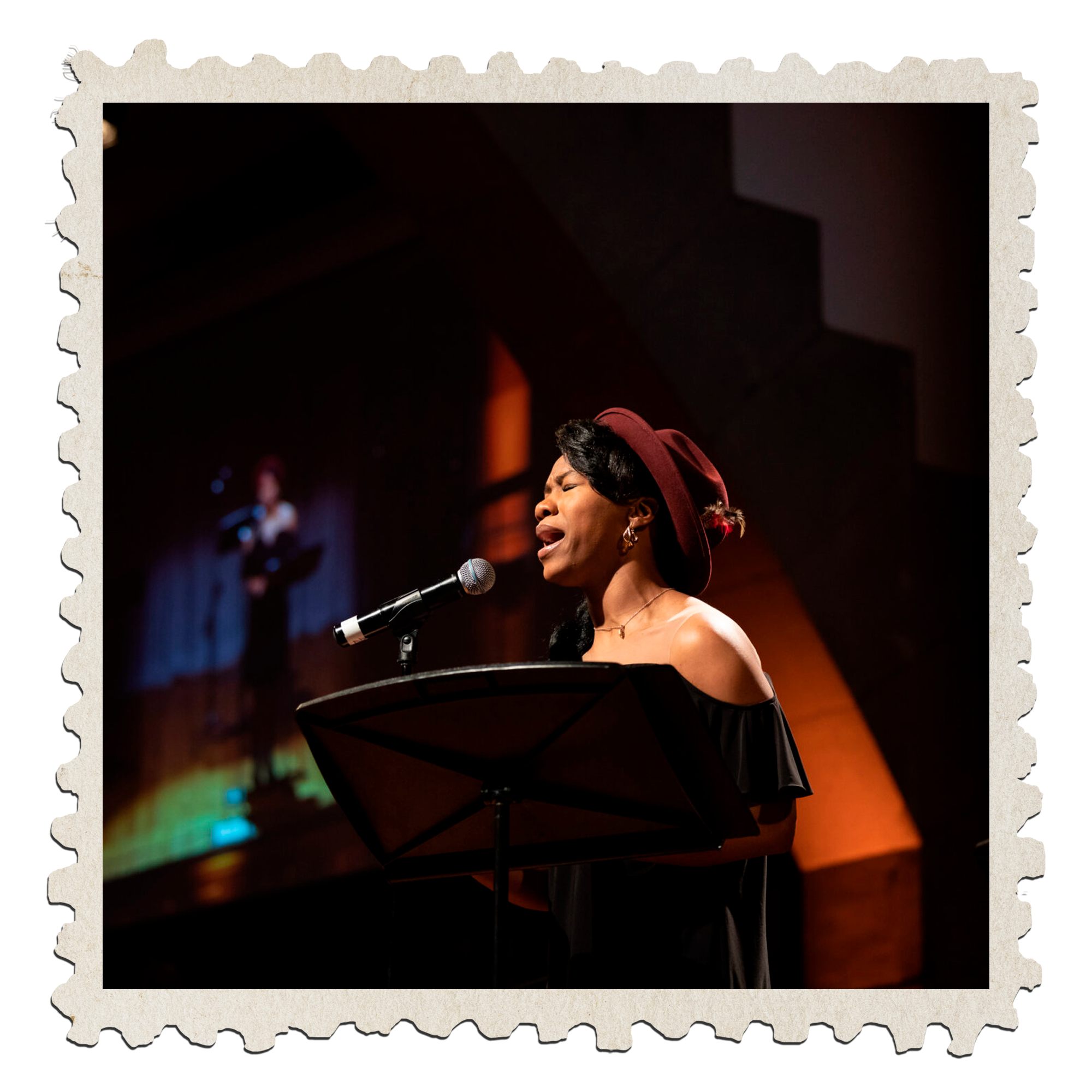 A woman with dark hair and earrings singing passionately at a microphone on stage, wearing a maroon hat and black dressed, in a performance setting with warm lighting.