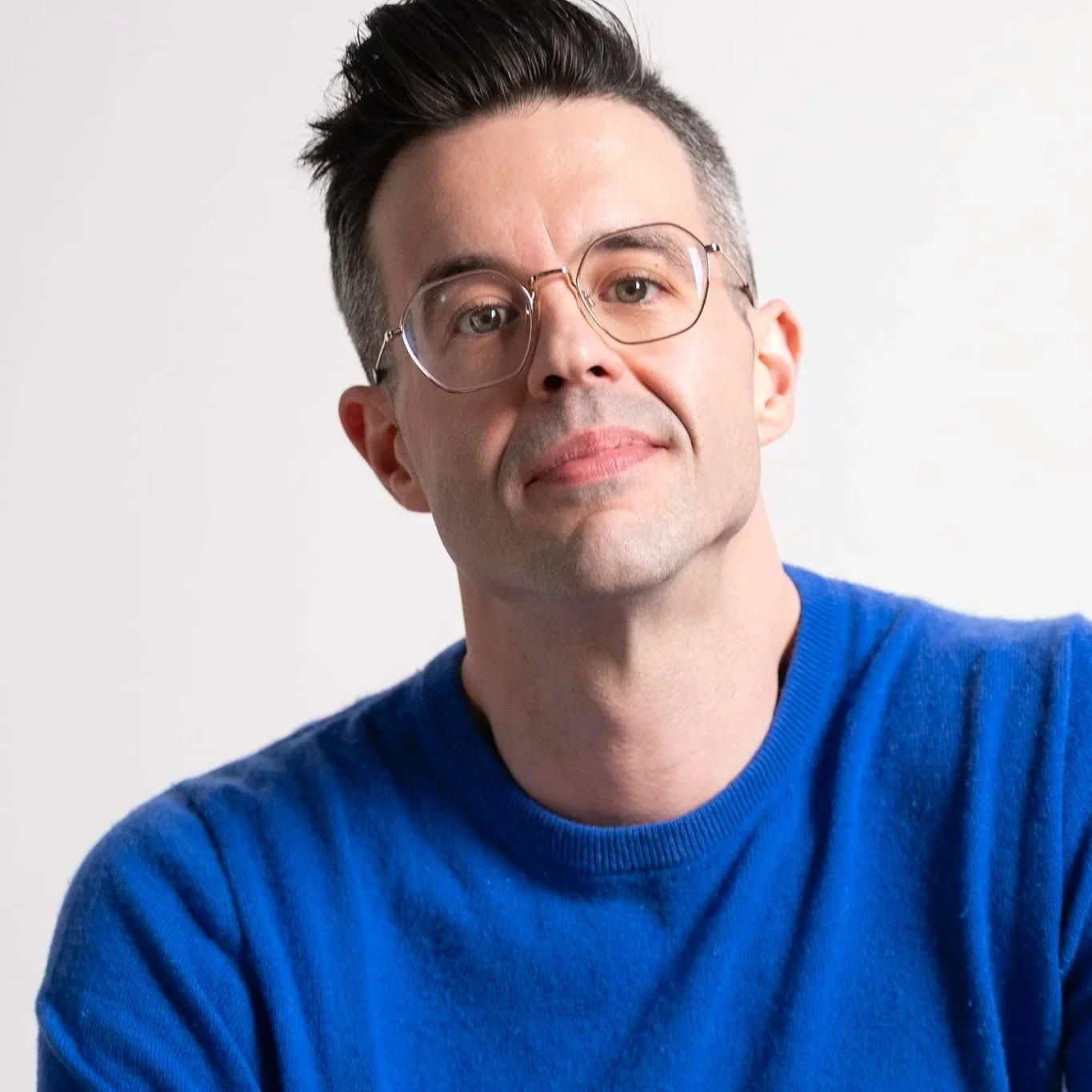 A man with dark hair, wearing glasses and a blue sweater, looking at the camera with a slight smile against a plain white background.