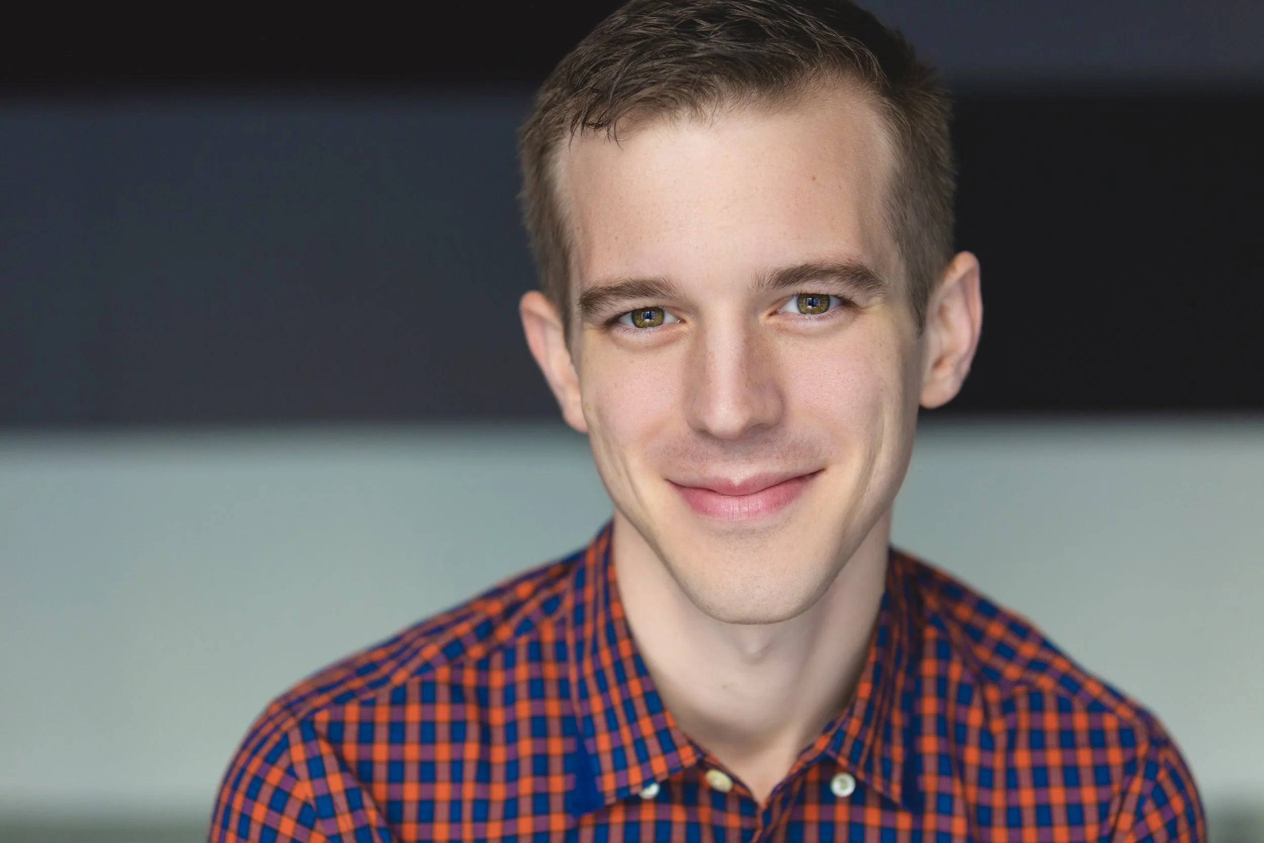 Close-up of a young man with light skin and short brown hair, wearing a red and blue plaid shirt, smiling at the camera.